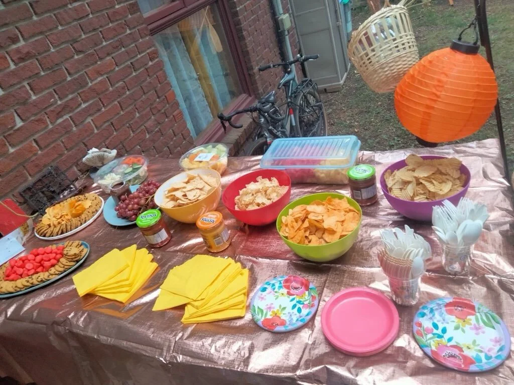 Outdoor party table with snacks, chips, dip, grapes, cookies, and paper plates, set against a brick wall with bicycles in the background, decorated with orange and white paper lanterns.