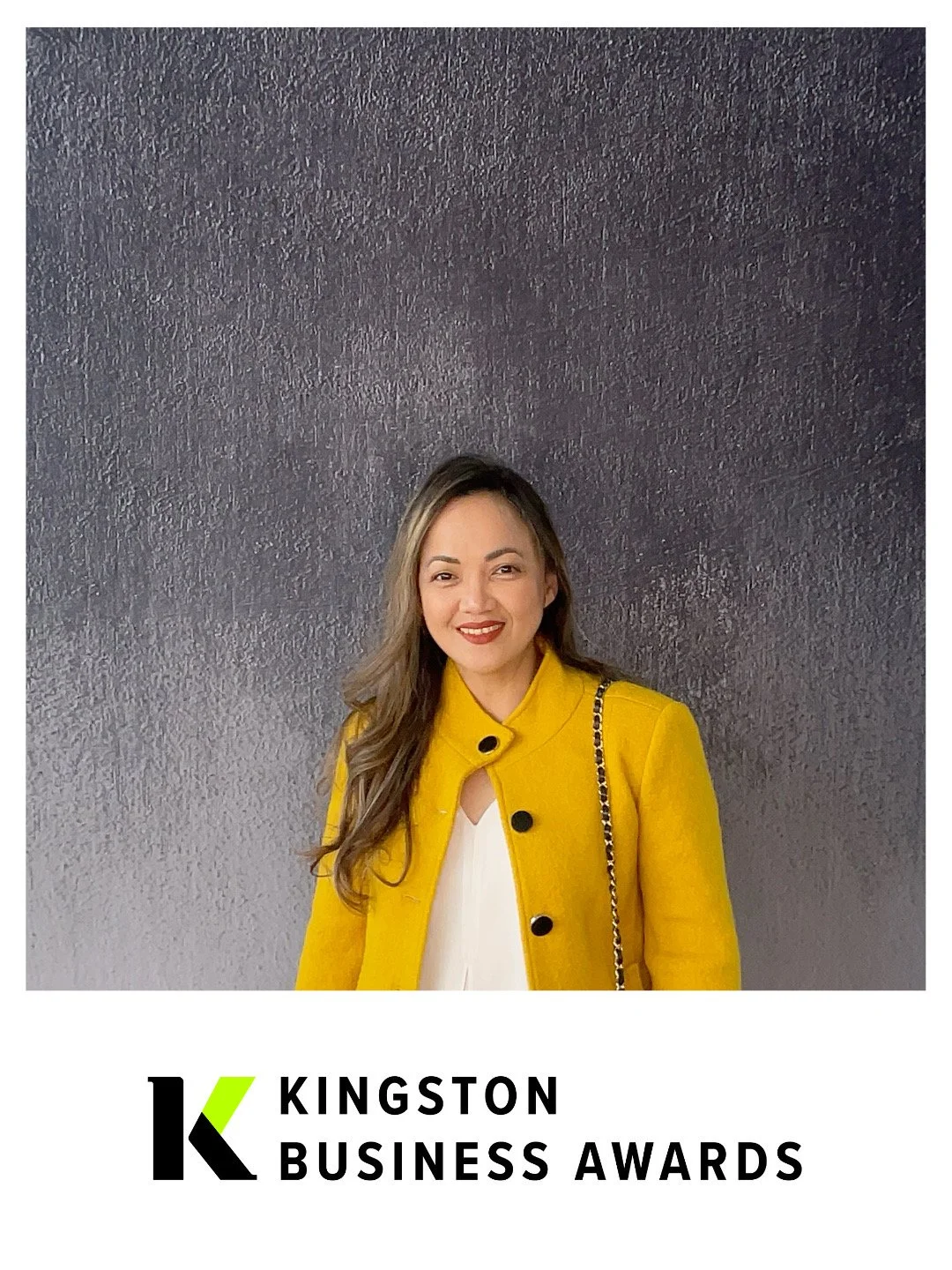 A woman with long wavy hair wearing a yellow coat with black buttons, standing against a dark textured background, smiling. The bottom of the image features the logo and text for Kingston Business Awards.