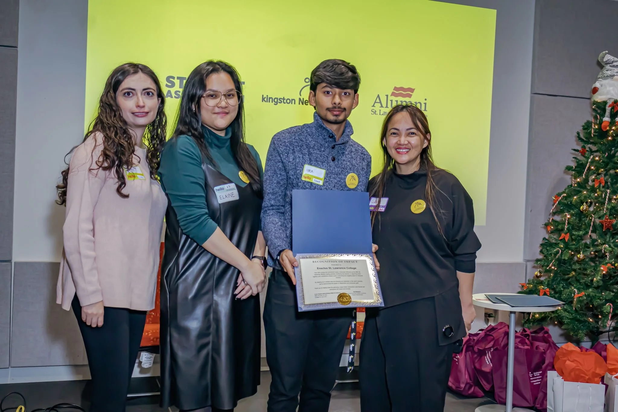 Group of five people standing in front of a yellow backdrop, one person holding a certificate, Christmas tree decorated with ornaments and ribbons on the right.