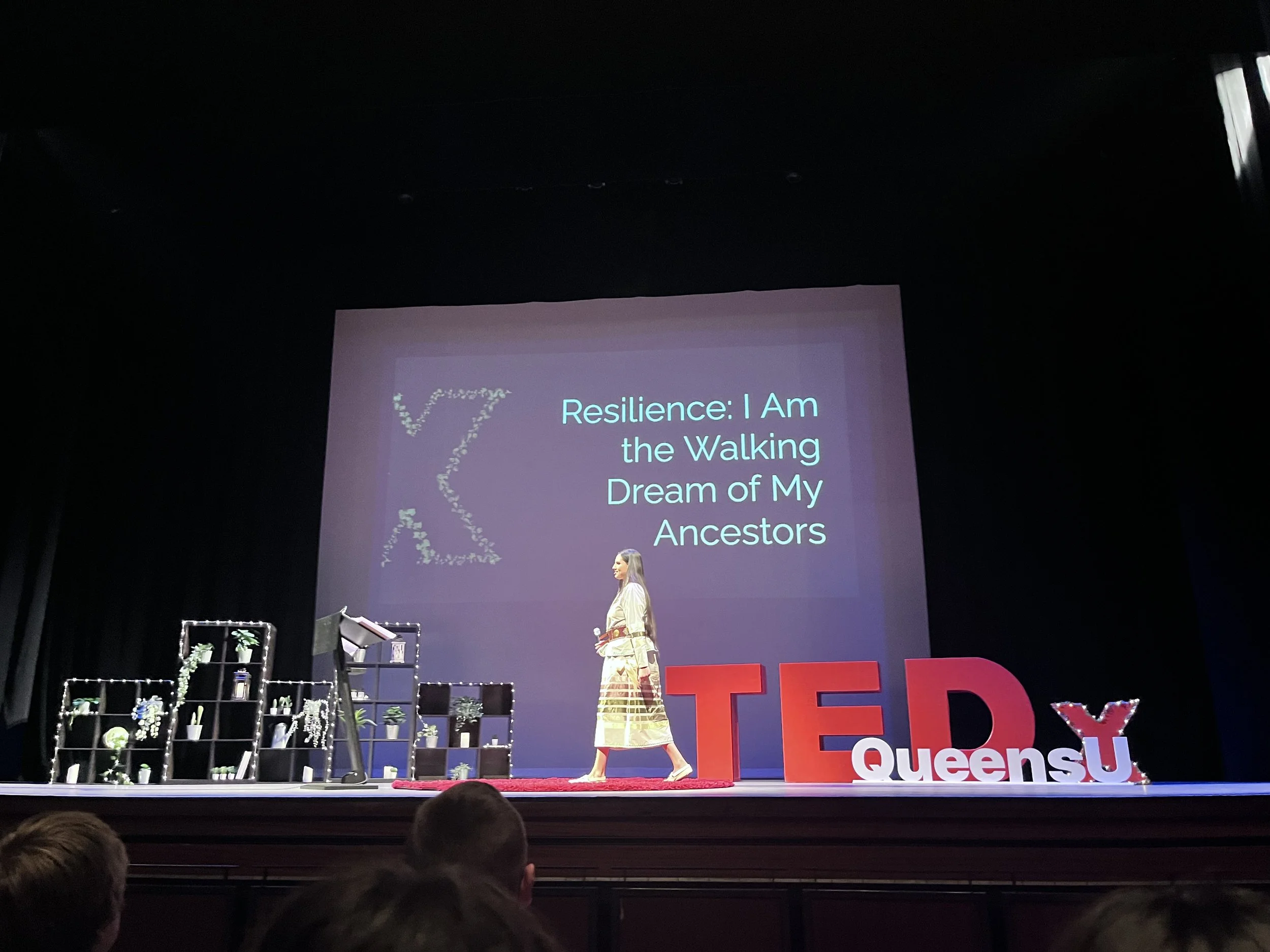 A woman standing on stage during a TEDxQueensU event, with a large screen behind her displaying the words 'Resilience: I Am the Walking Dream of My Ancestors' and decorated large red letters spelling 'TED' and smaller white letters spelling 'QueensU'.