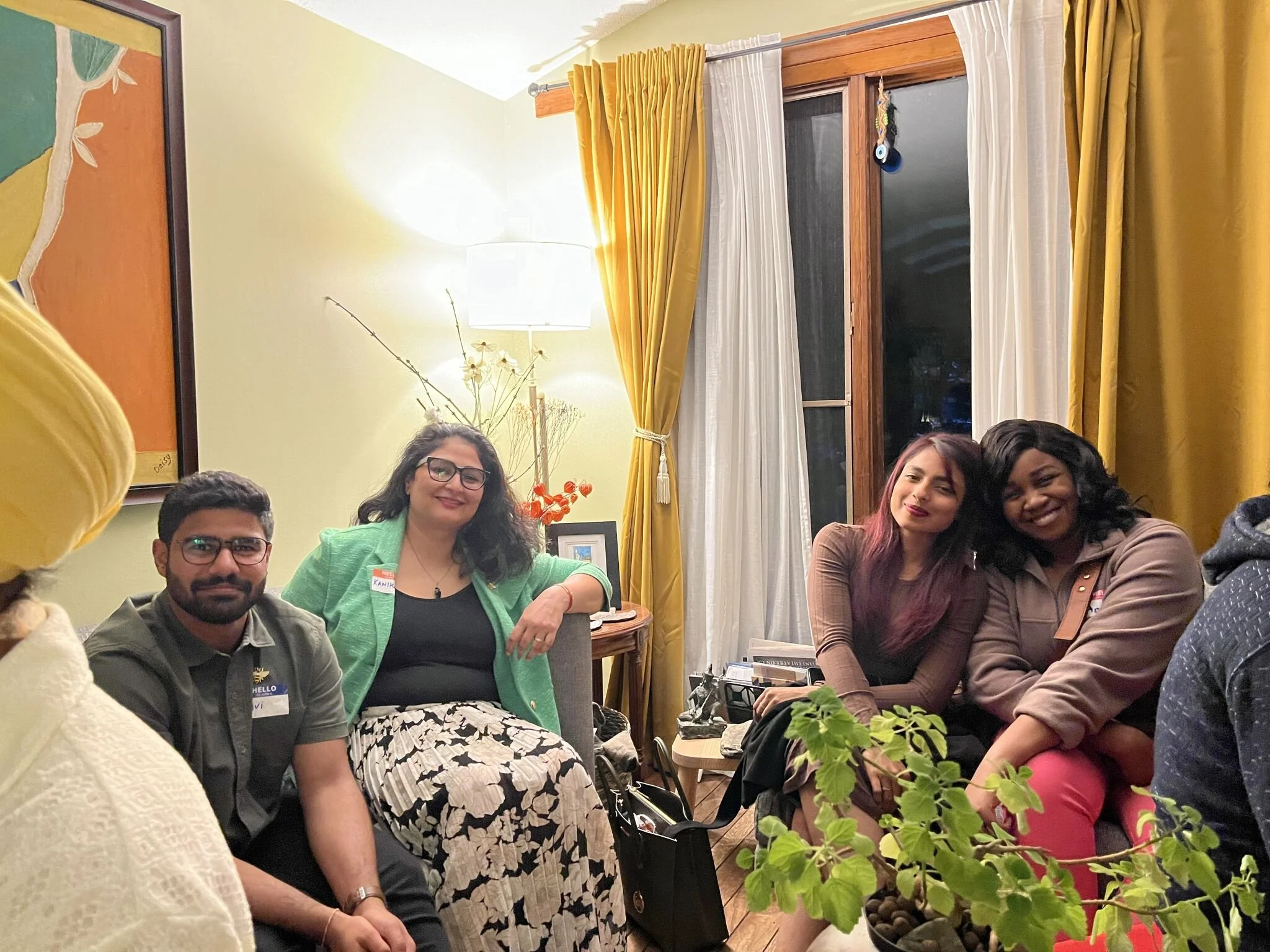 Four people sitting comfortably in a cozy living room, smiling for the camera. The room features yellow curtains, a window, and a table with decorative items, including a vase with flowers and a framed photo.
