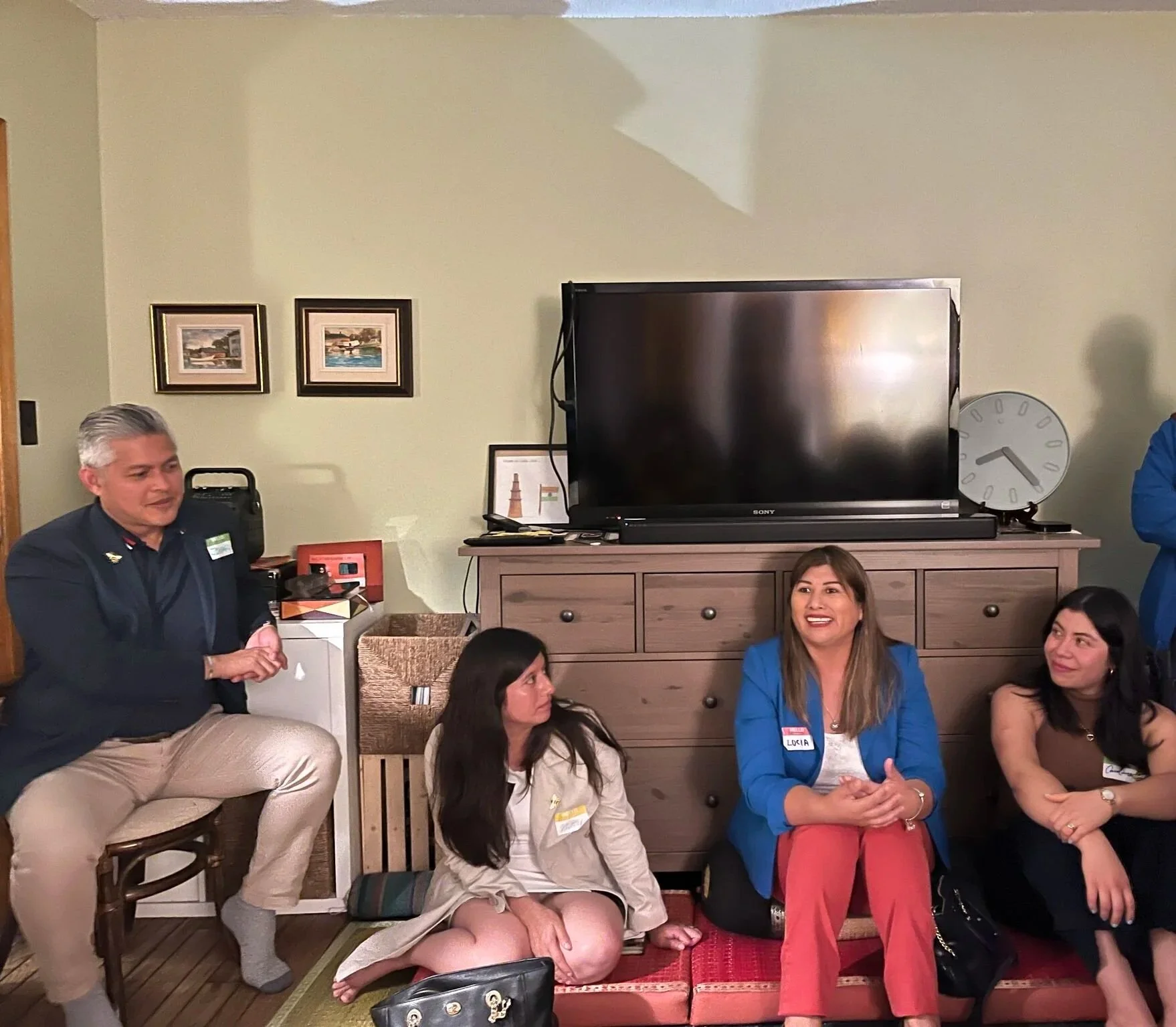 Group of people sitting on the floor and a small stool in a room, engaged in conversation. There are three women sitting on cushions, and a man sitting on a small stool. Behind them is a wooden cabinet with a large flat-screen TV, a clock, and framed pictures on the wall.