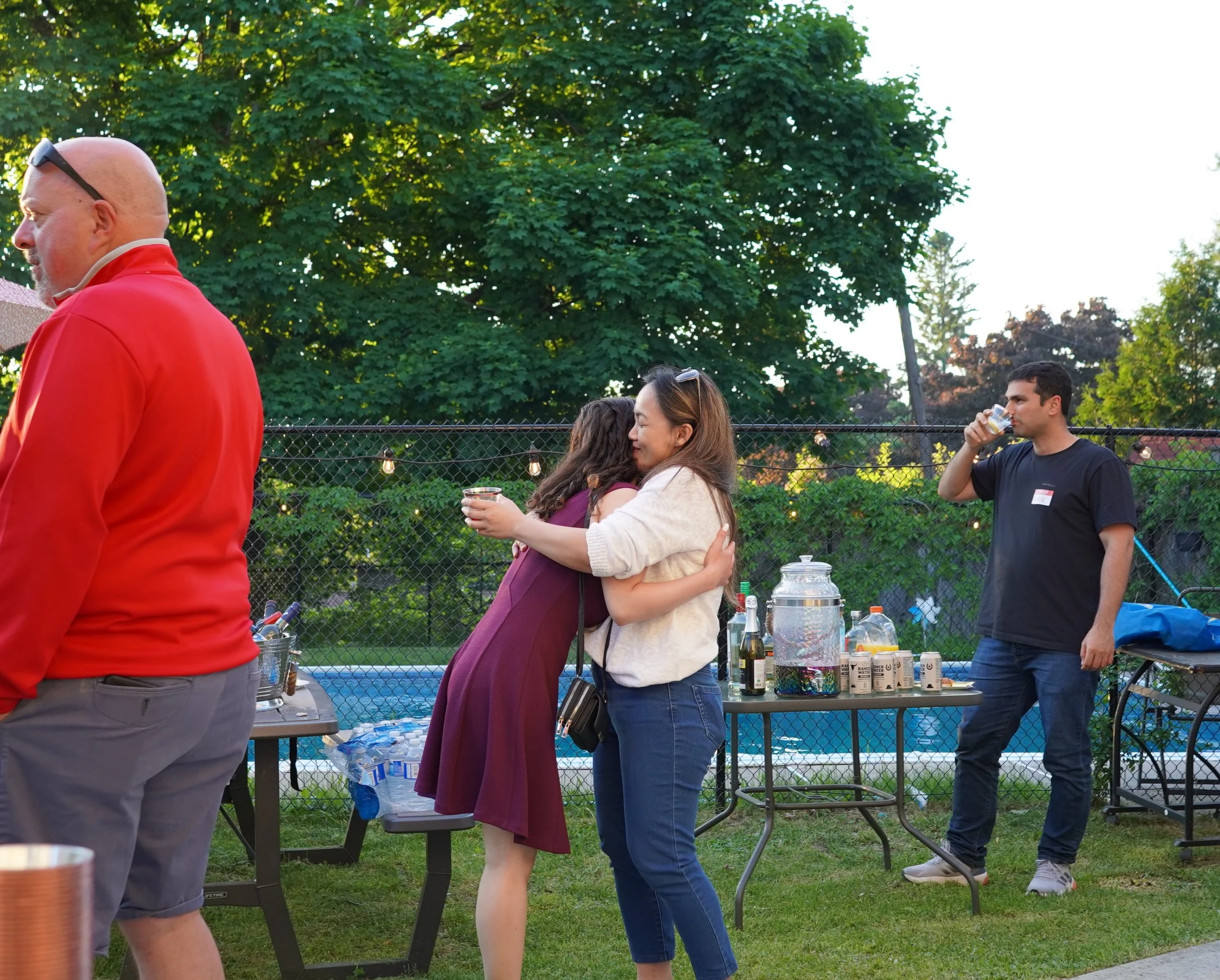 People at a backyard gathering, hugging, drinking, and standing near a table with beverages and snacks, with a pool and trees in the background.