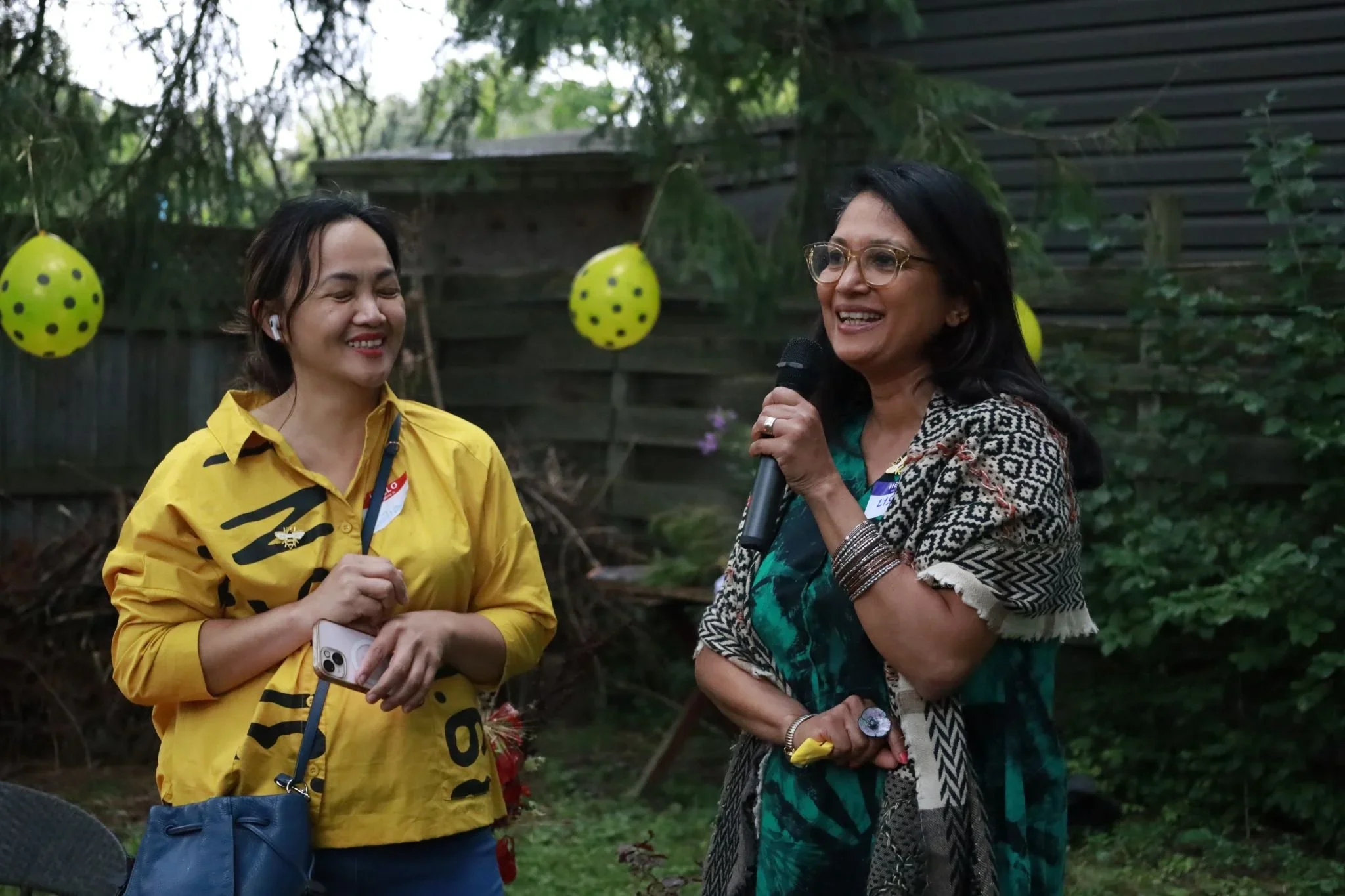 Two women at an outdoor celebration, one holding a microphone and the other smiling, with yellow and black polka-dotted balloons hanging in the background.