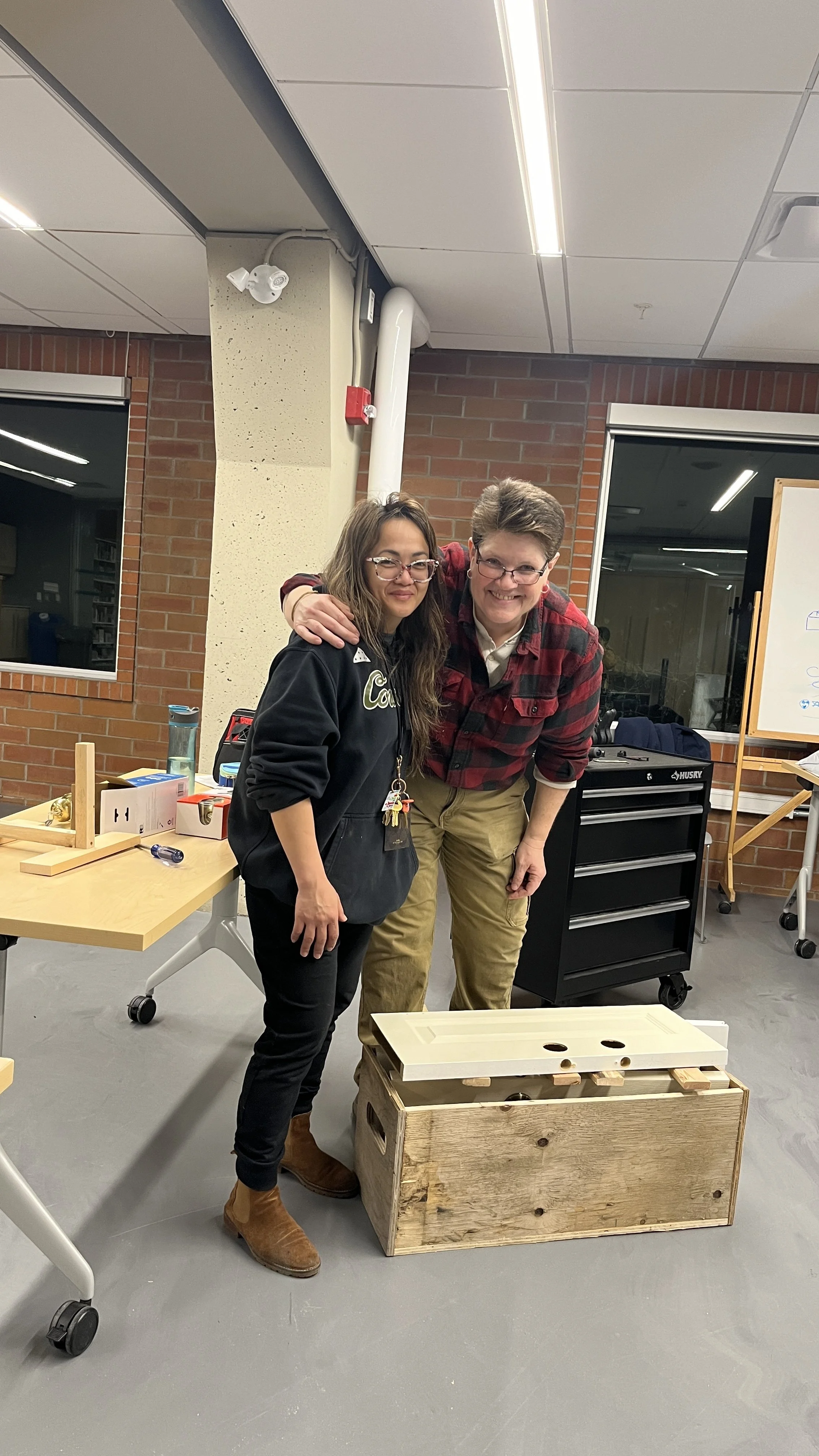Two people crouch next to a wooden crate in a room, smiling at the camera. One wears a hoodie and glasses, the other a plaid shirt and glasses. There is a whiteboard, a black tool chest, and tables with various objects in the background.