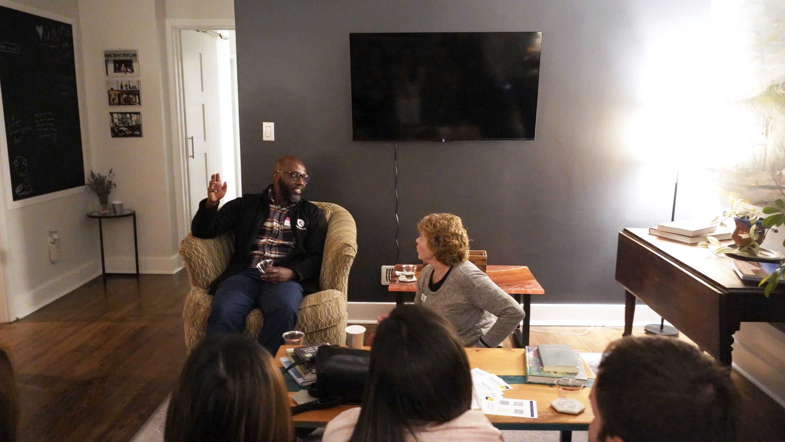 A man and a woman are sitting and talking in a living room, with two onlookers in the foreground. The man is sitting on an armchair, raising one hand as he speaks. The woman is sitting on a small side table, listening. A large black TV is mounted on the dark gray wall behind them. There is a wooden console with books and a plant on it to the right. The room has hardwood floors and a side table with flowers near the doorway.