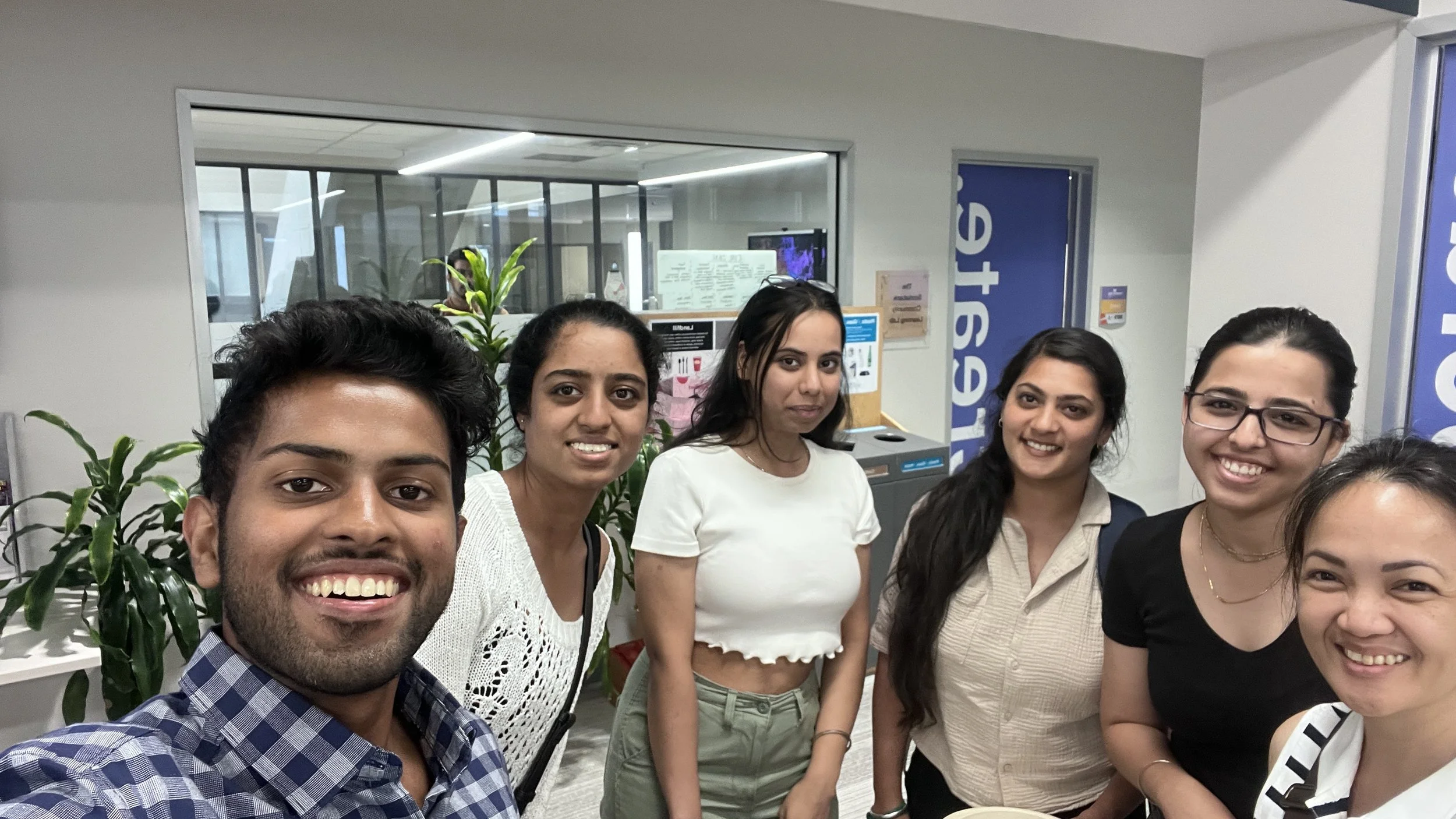 Group of six diverse people smiling and taking a selfie indoors, with plant and office background.
