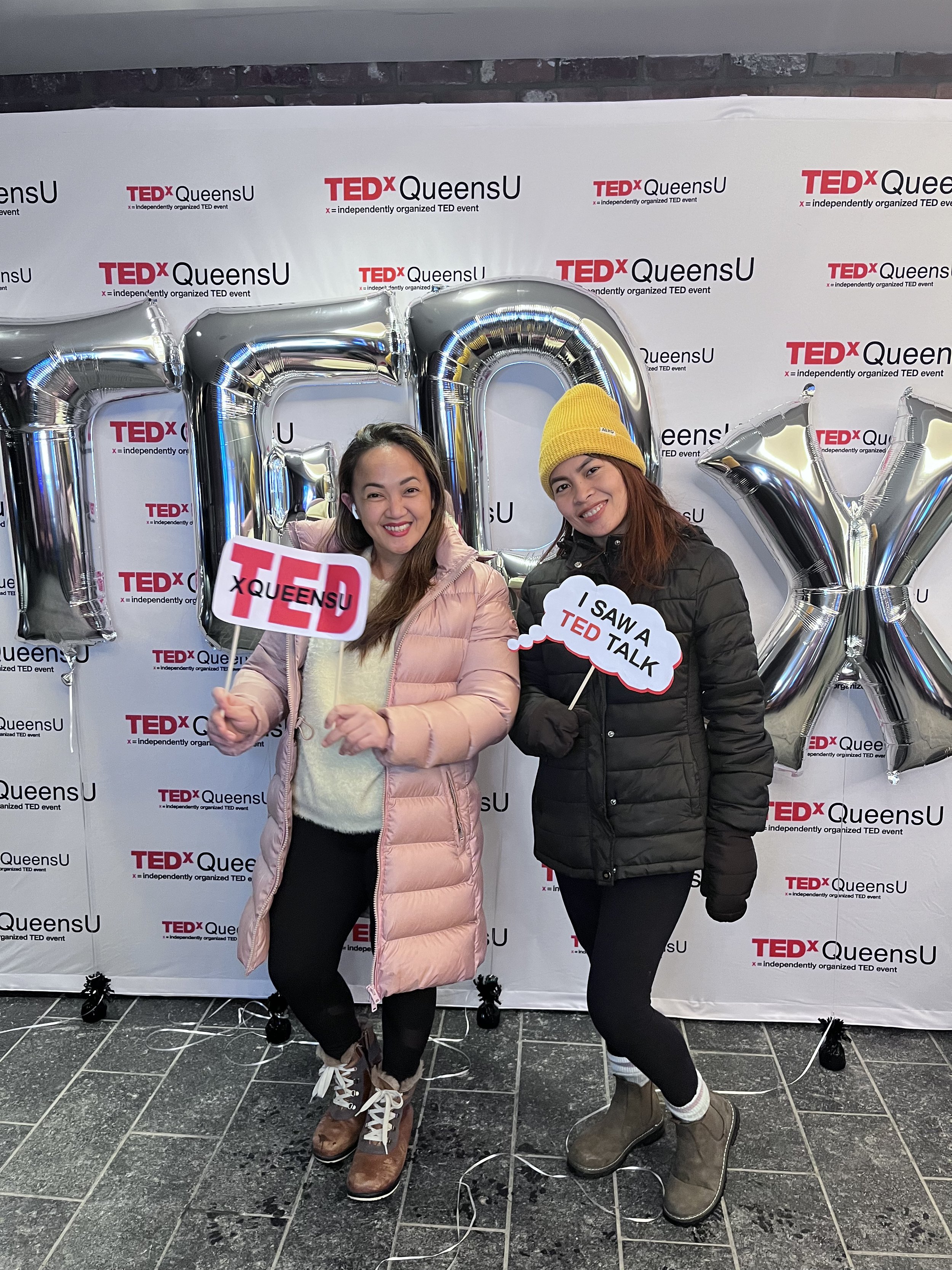 Two women at a TEDx event in QueensU, standing in front of a branded backdrop with large silver balloons spelling 'TED' in the background. They hold signs related to TED talks, smiling and dressed warmly.