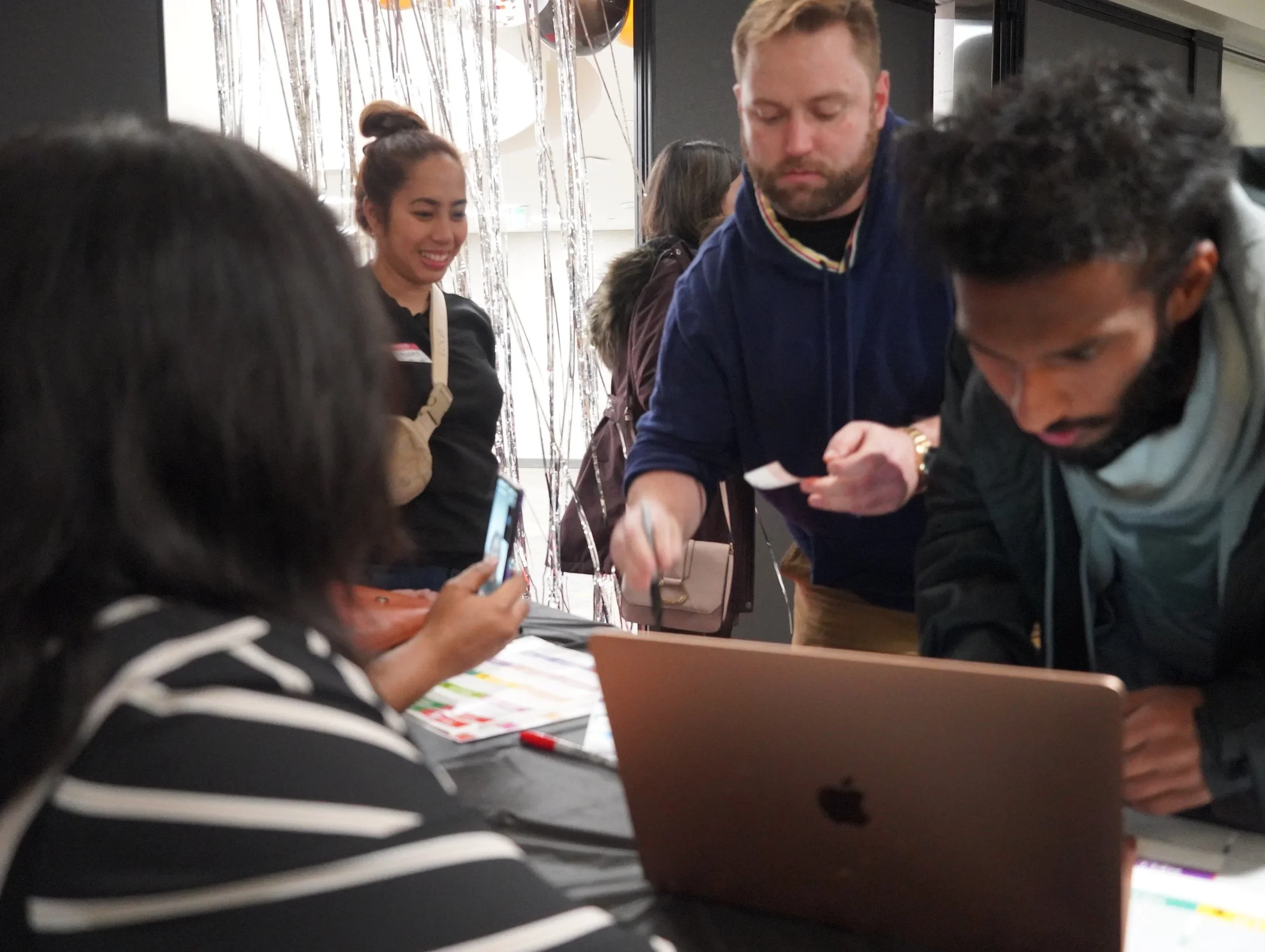 Group of people gathered around a table with a laptop, engaged in discussion or activity, with a woman taking a photo with her smartphone, and a woman smiling in the background.