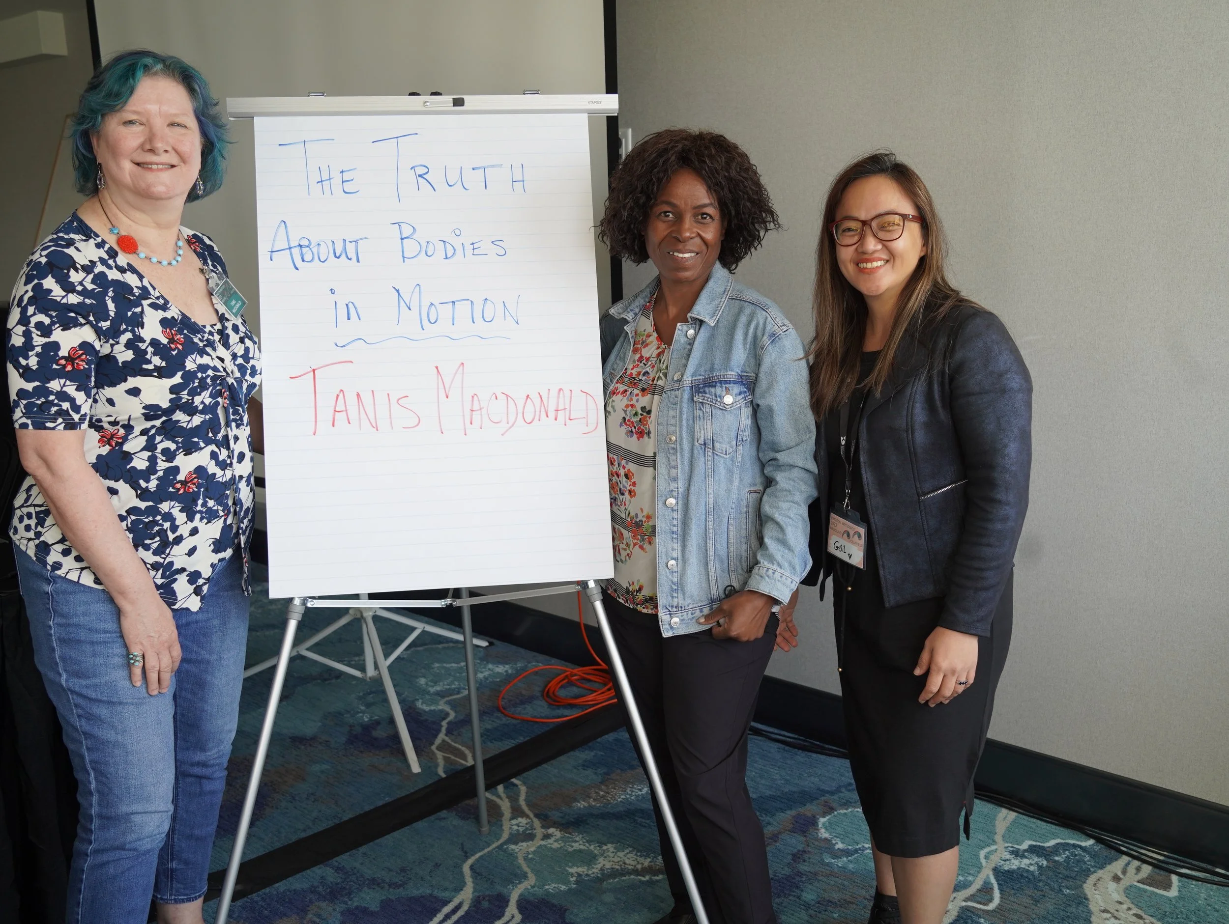 Three women standing next to a whiteboard with a handwritten presentation title, posing for a photo in an indoor conference room.