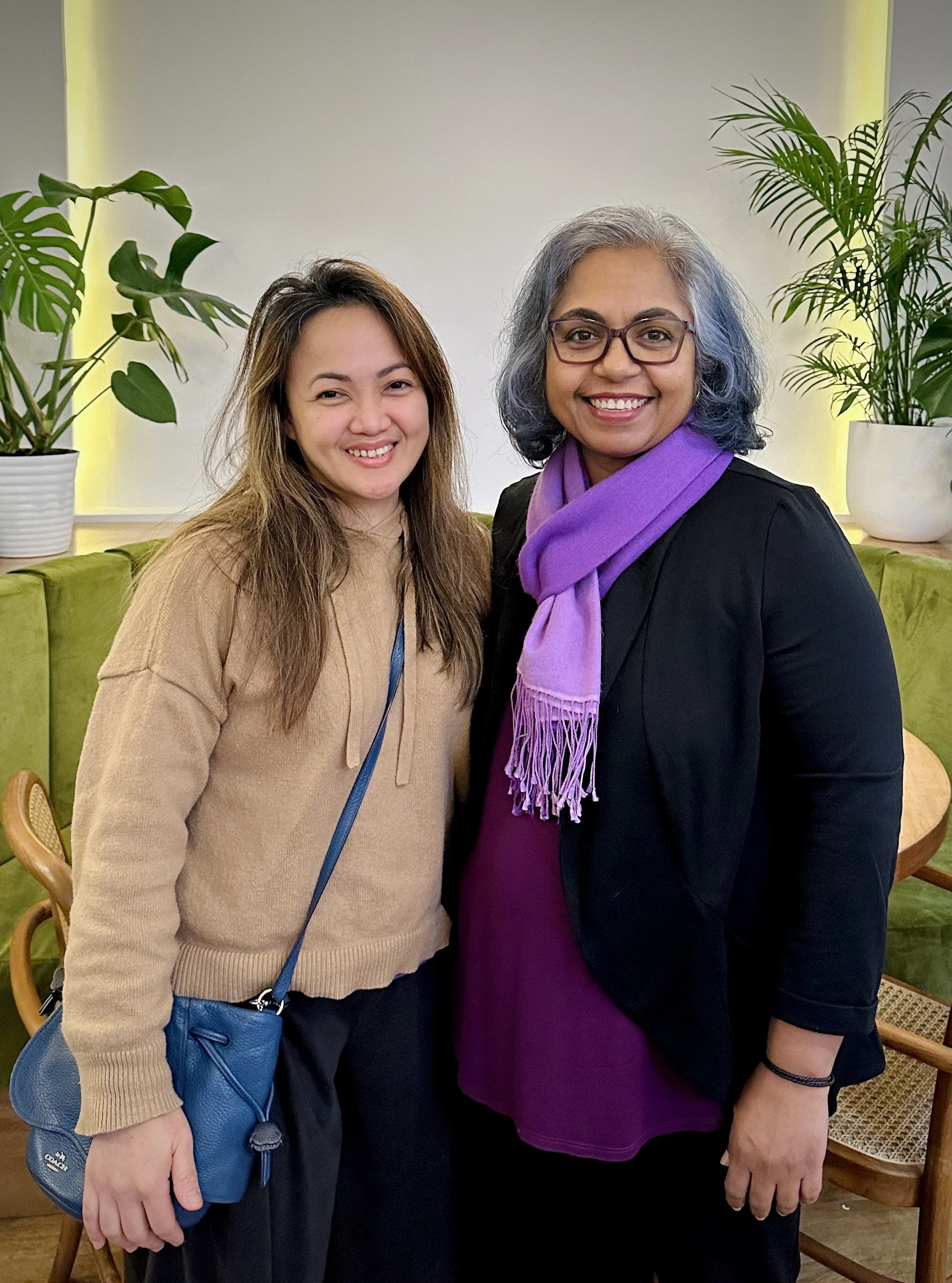 Two women standing together in a room with green plants, smiling at the camera.