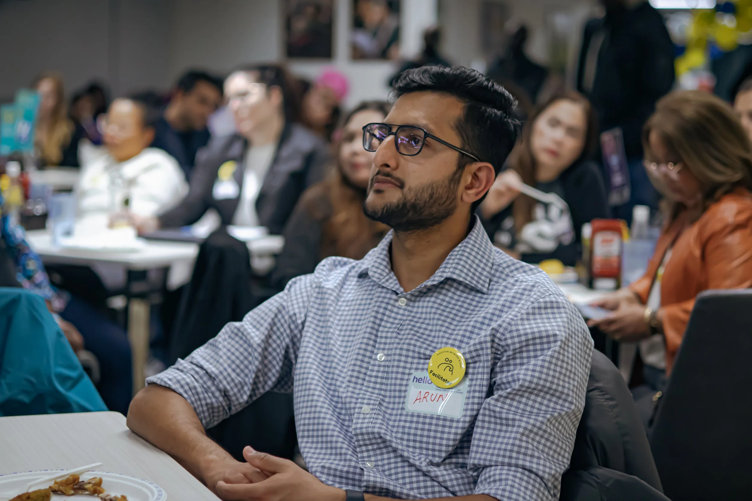A man with glasses and a checkered shirt, wearing a name tag with "Arun" and a yellow "Facilitator" badge, is attending a conference or seminar in a room filled with other people sitting at tables.