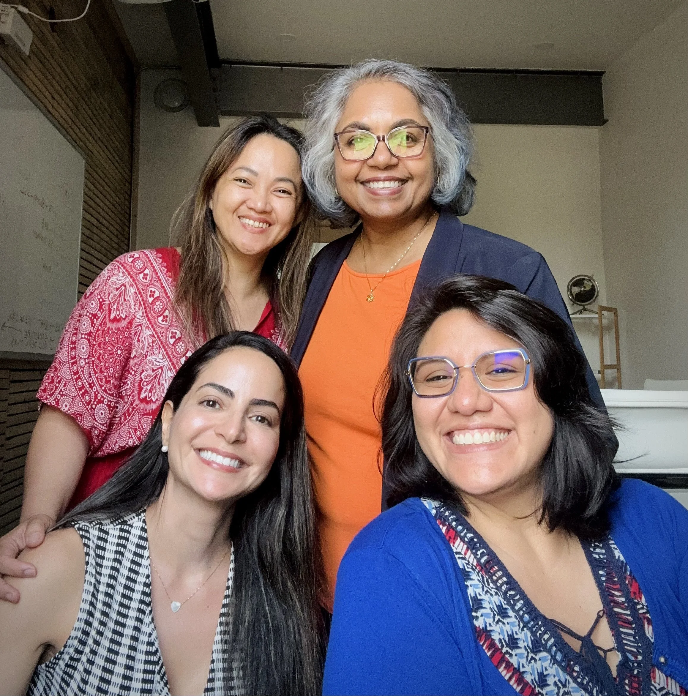 Group of five women smiling indoors, with diverse appearances, two sitting and three standing.