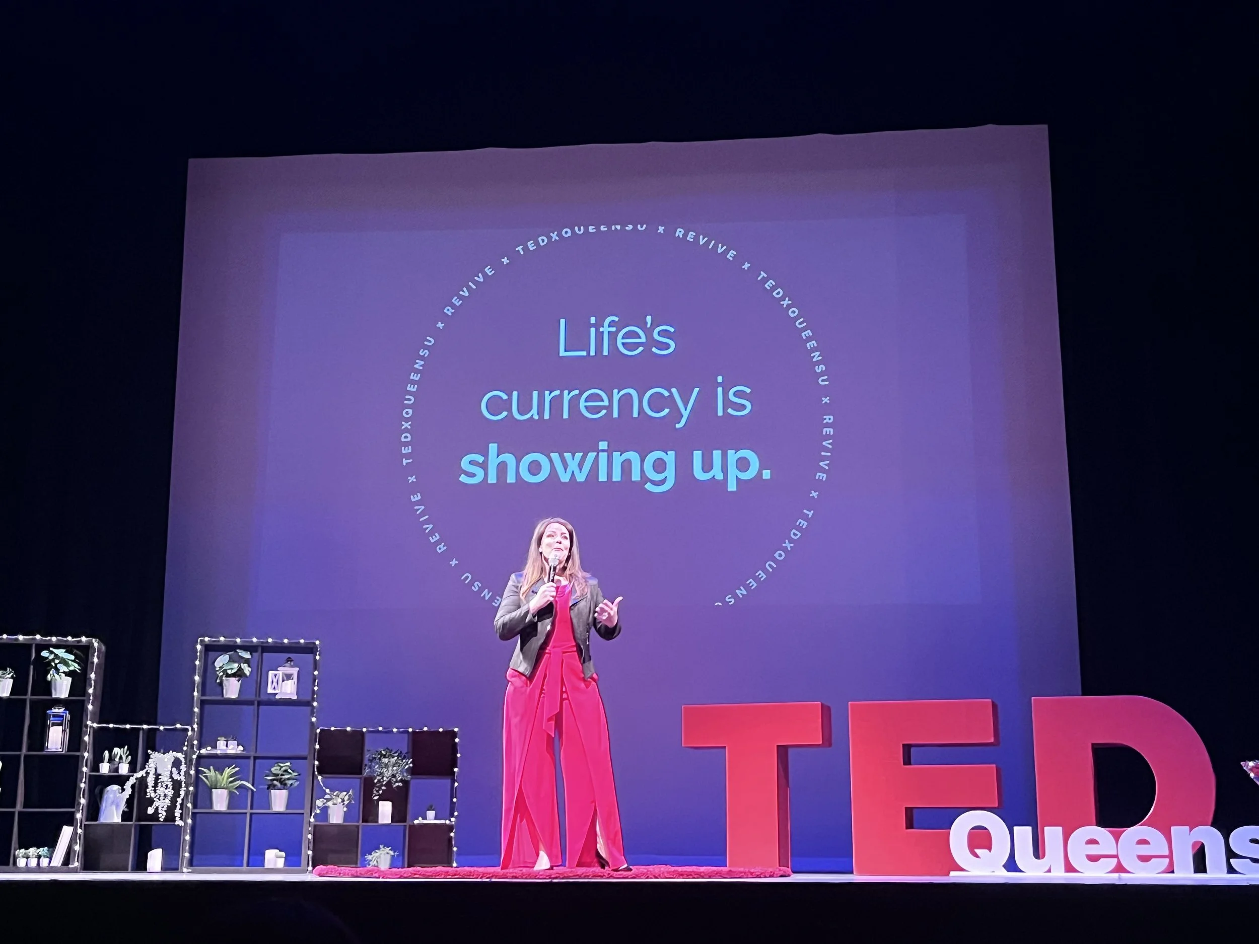 A woman standing on stage giving a presentation at a TEDx event. The background screen displays the quote 'Life's currency is showing up.' The stage has shelves with plants and decor, and large red and white letters spell out 'TED Queens'.