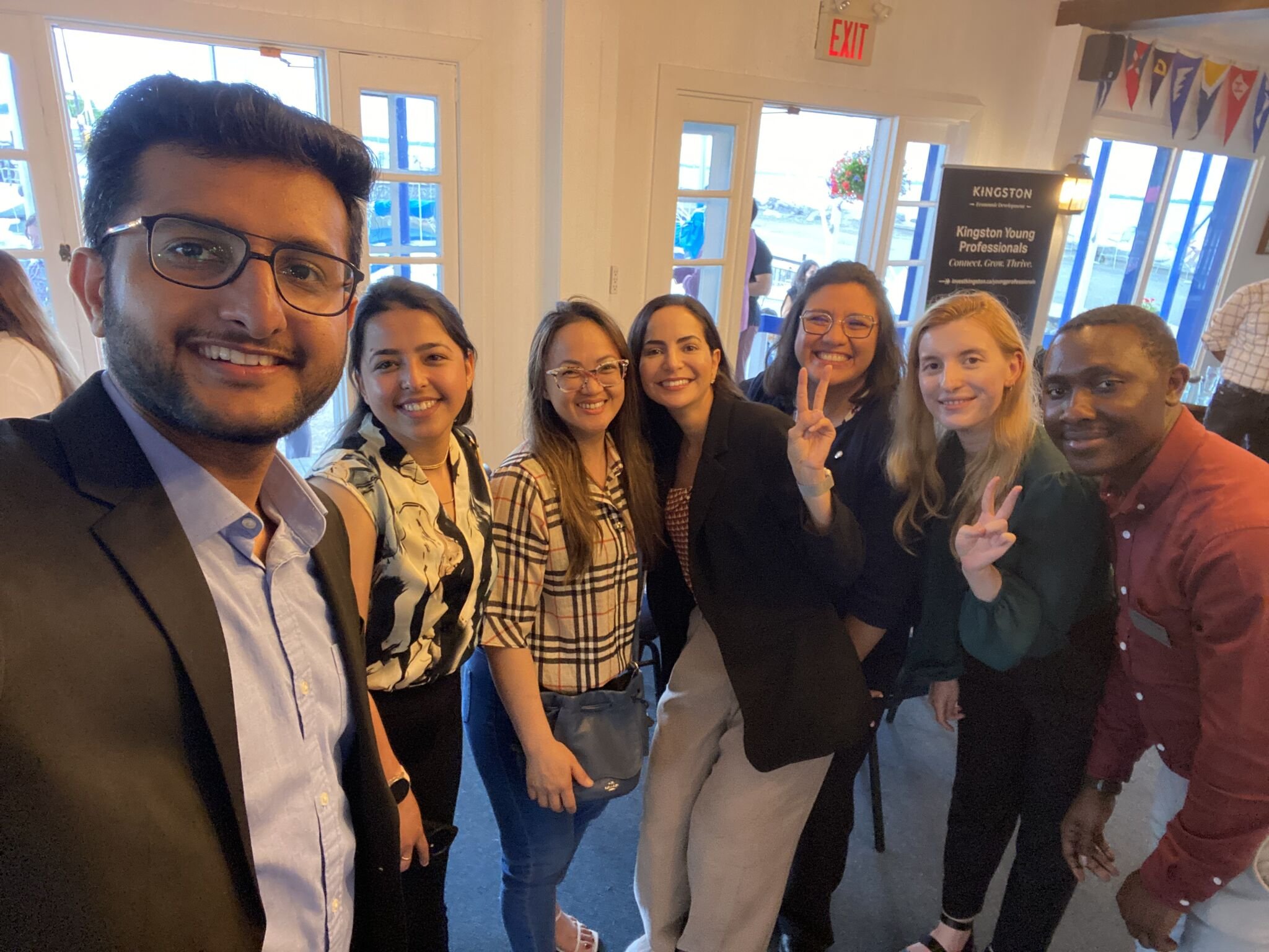 Group of seven smiling young adults taking a selfie indoors, with windows and an exit sign in the background.