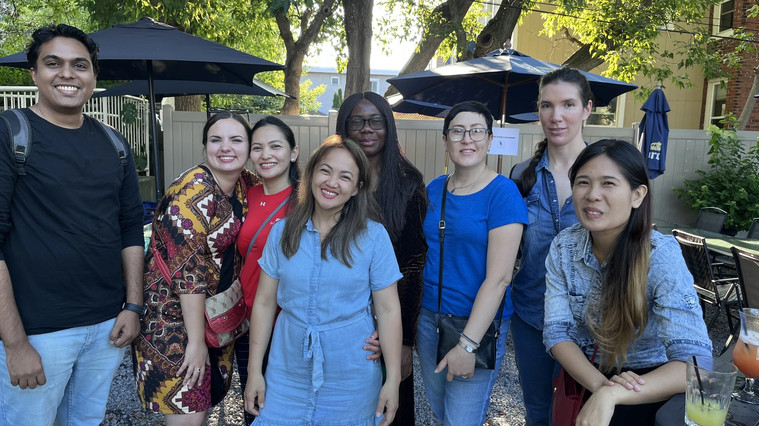 Group of eight diverse people smiling outdoors at a gathering with umbrellas, trees, and a fenced backyard in the background.