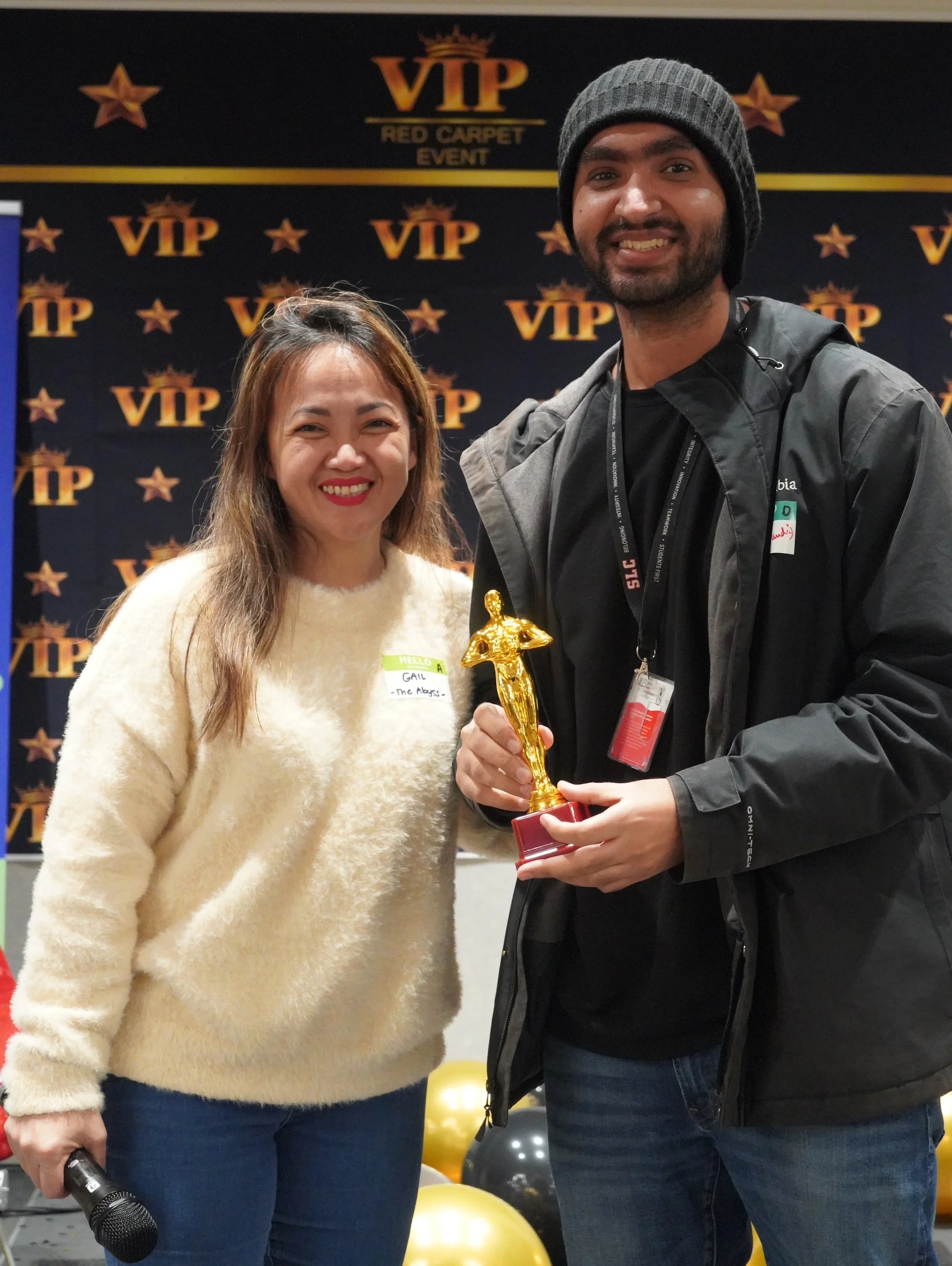 A woman and a man smiling at an event, with the woman holding a microphone and the man holding a gold award trophy shaped like an Oscar. The background displays a step and repeat banner with the VIP logo and stars, indicating a VIP red carpet event.