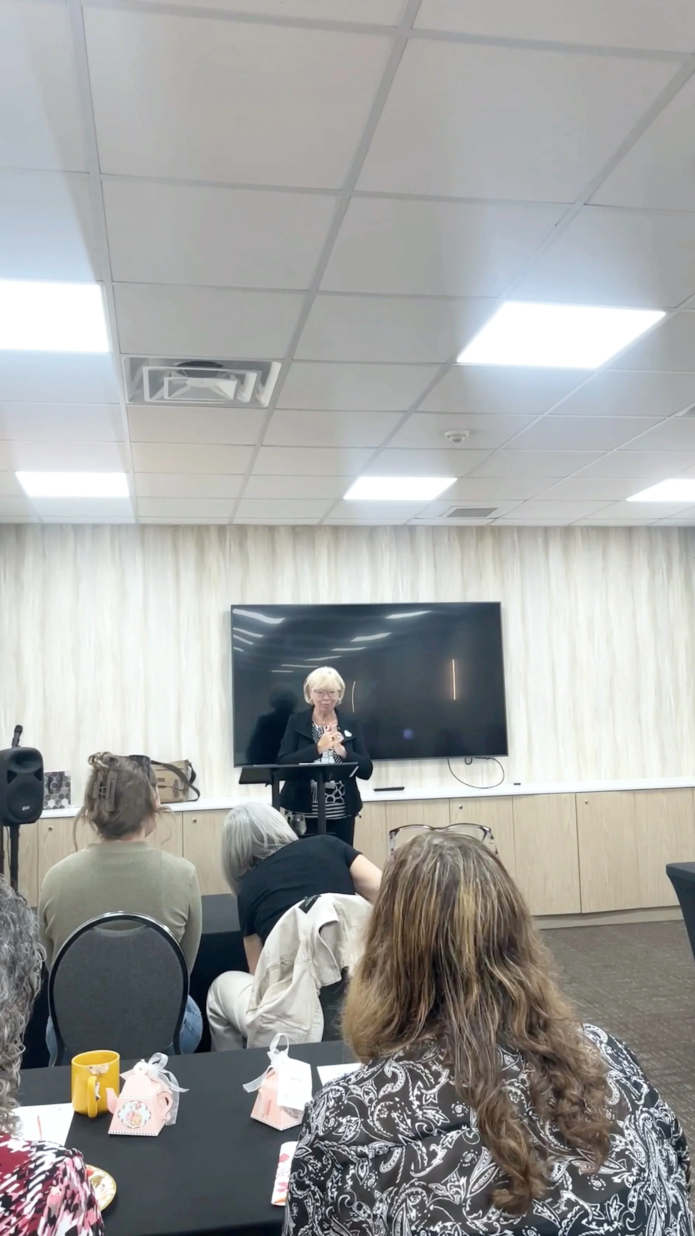 A woman standing at a podium giving a presentation in front of a black television screen, with several attendees seated at tables in a conference room.