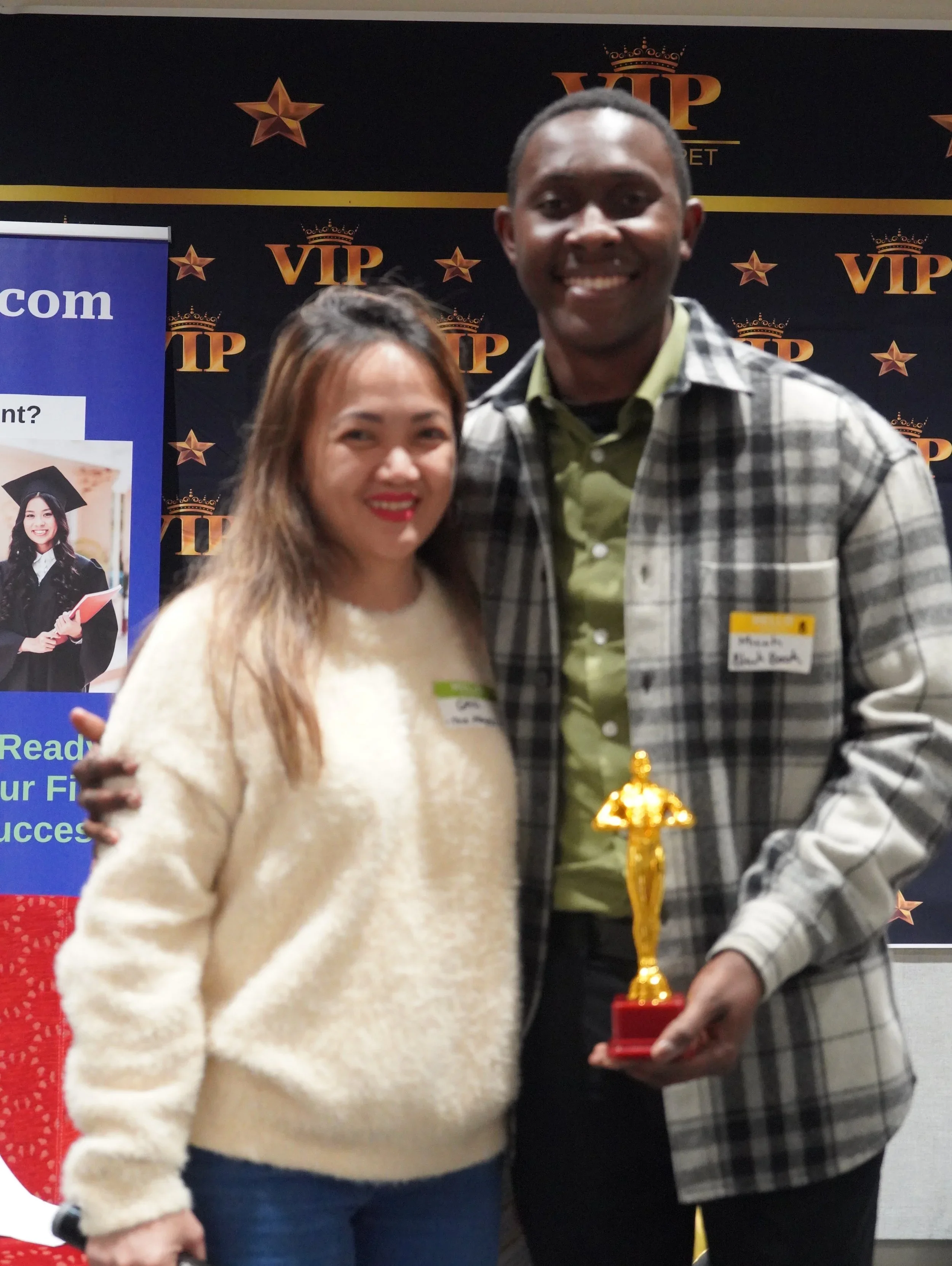 A woman and a man smiling, standing together at an awards event, holding a small golden trophy. The woman has long brown hair, wearing a cream-colored sweater, and the man has short dark hair, wearing a green shirt and checkered jacket. Behind them is a black backdrop with golden stars and the word 'VIP'.