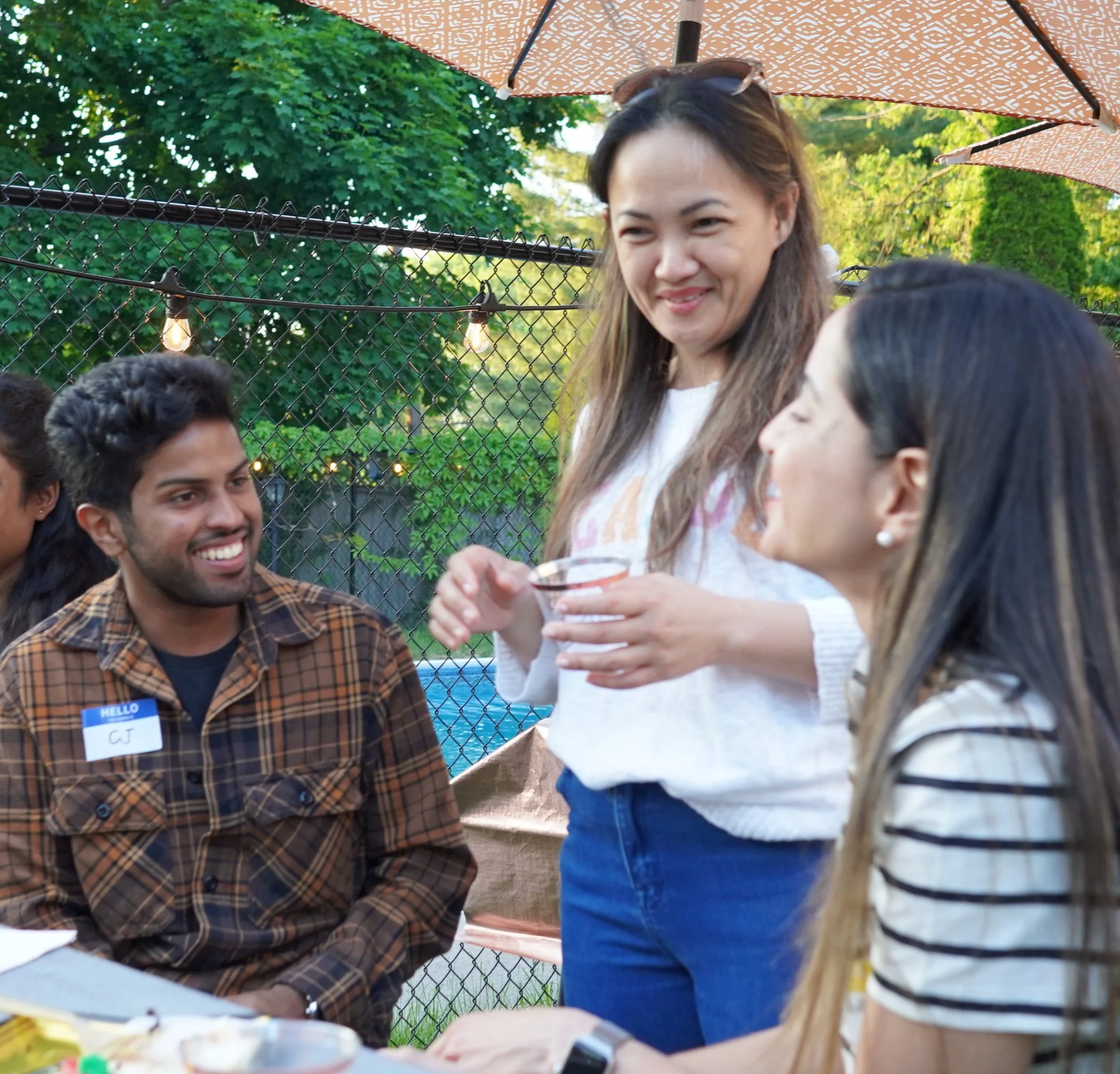 Group of diverse friends socializing outdoors under a large umbrella, with string lights and green trees in the background.