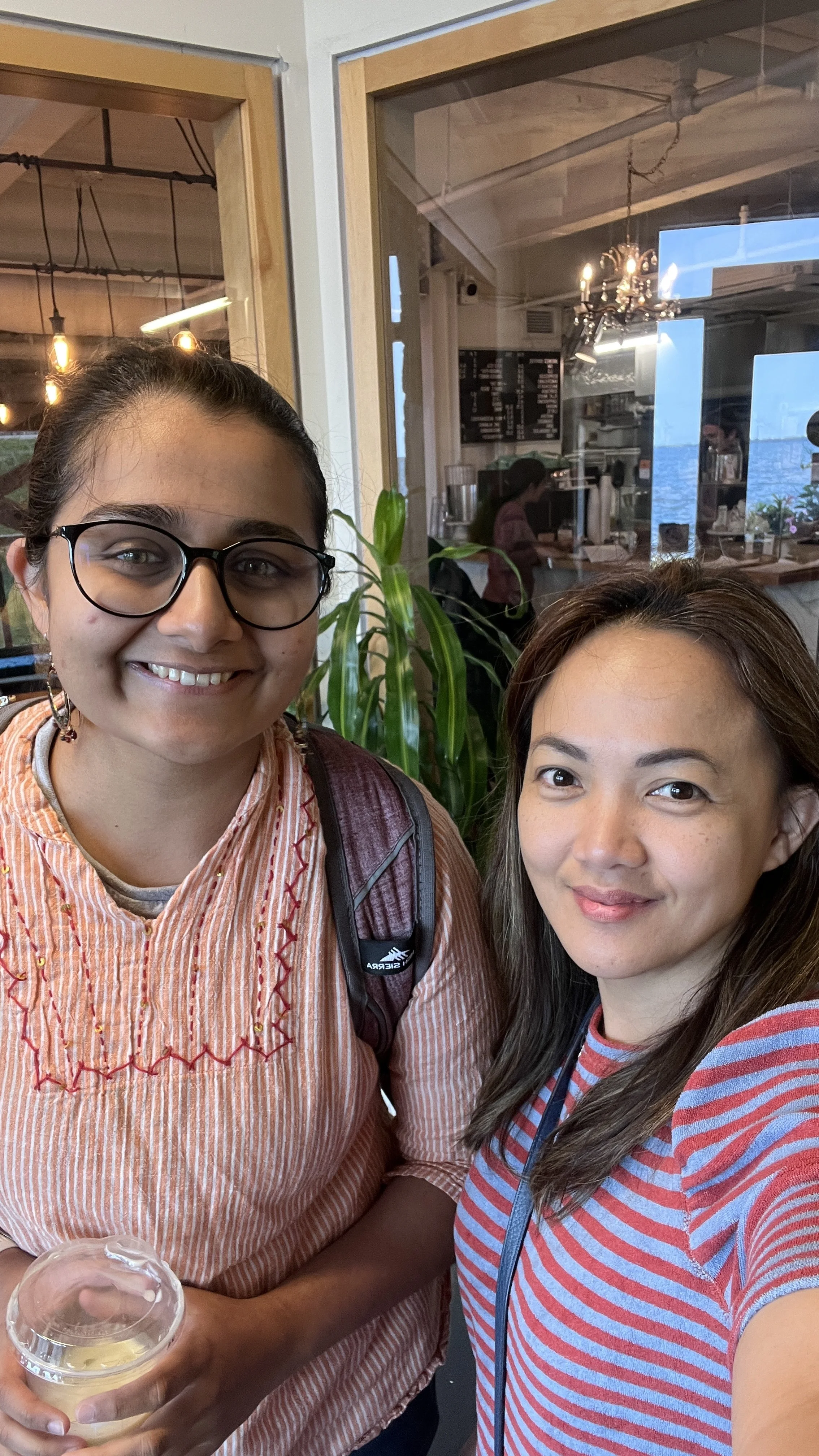 Two women taking a selfie inside a cafe or restaurant, with large windows behind them showing the interior and some customers at the counter.