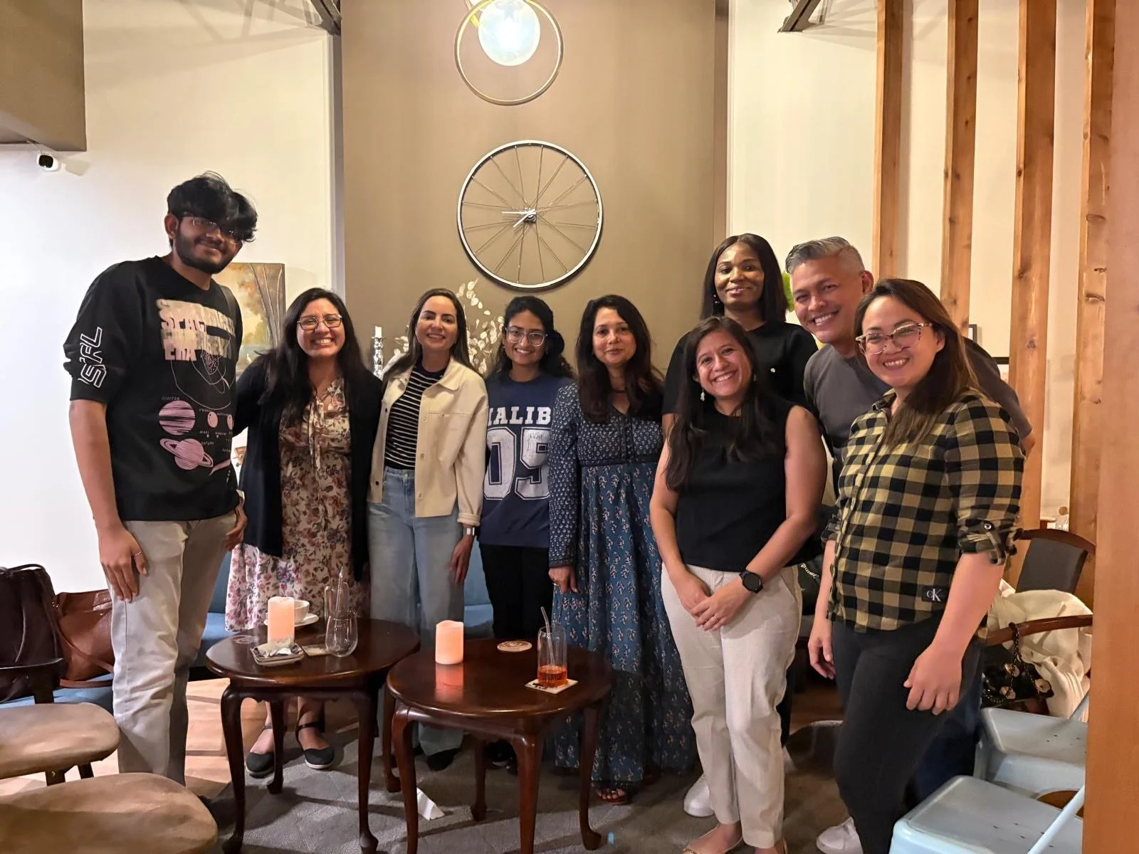 A group of nine people smiling and standing together indoors in a cozy room, with a wall clock and bicycle wheel decor behind them.