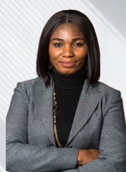 Portrait of a woman with shoulder-length black hair, wearing a grey blazer over a black turtleneck, standing against a light, patterned background.