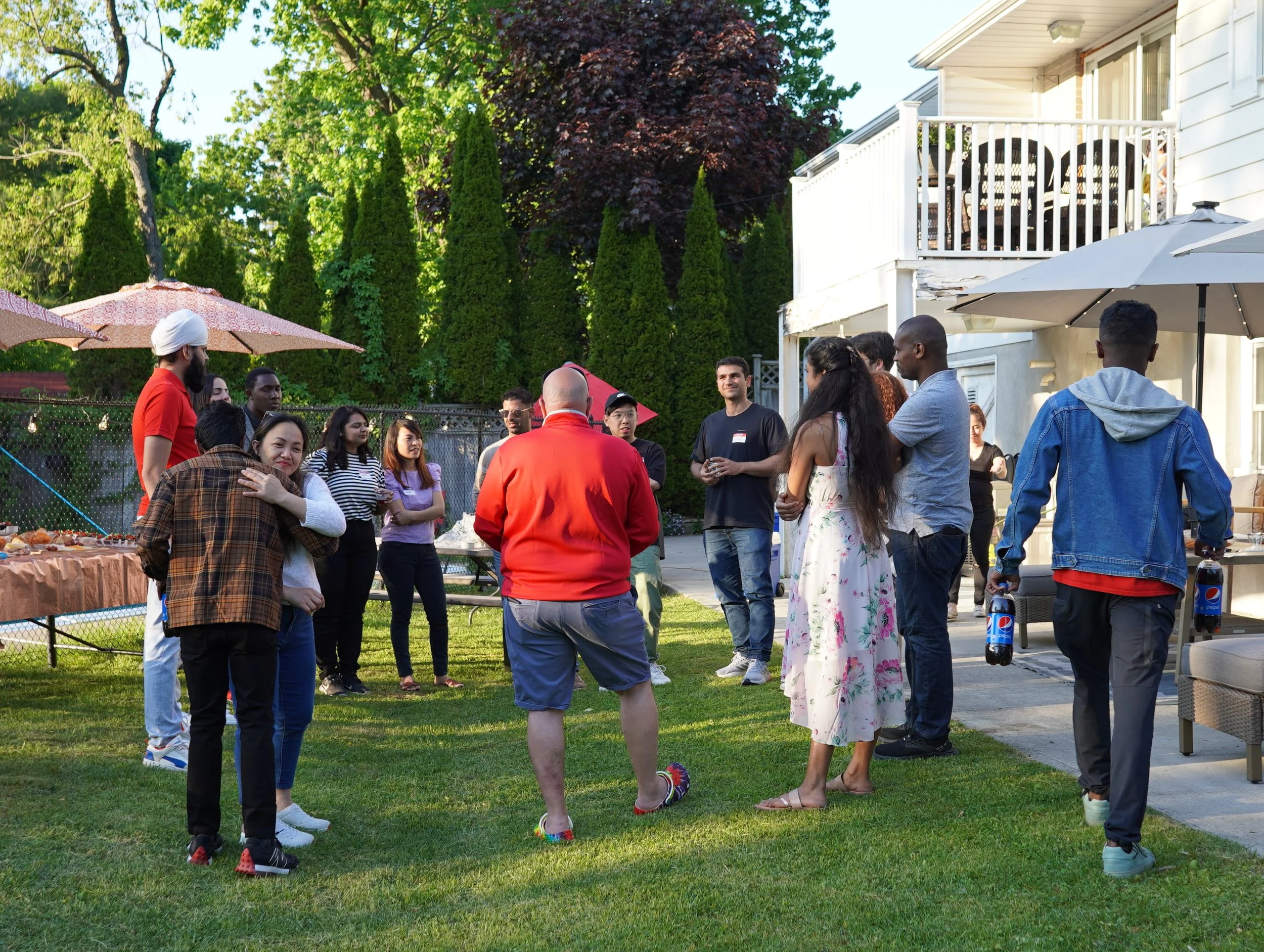 Group of people gathered outdoors for a social event, standing on a lawn with a white house in the background, some holding drinks, with umbrellas and trees around.