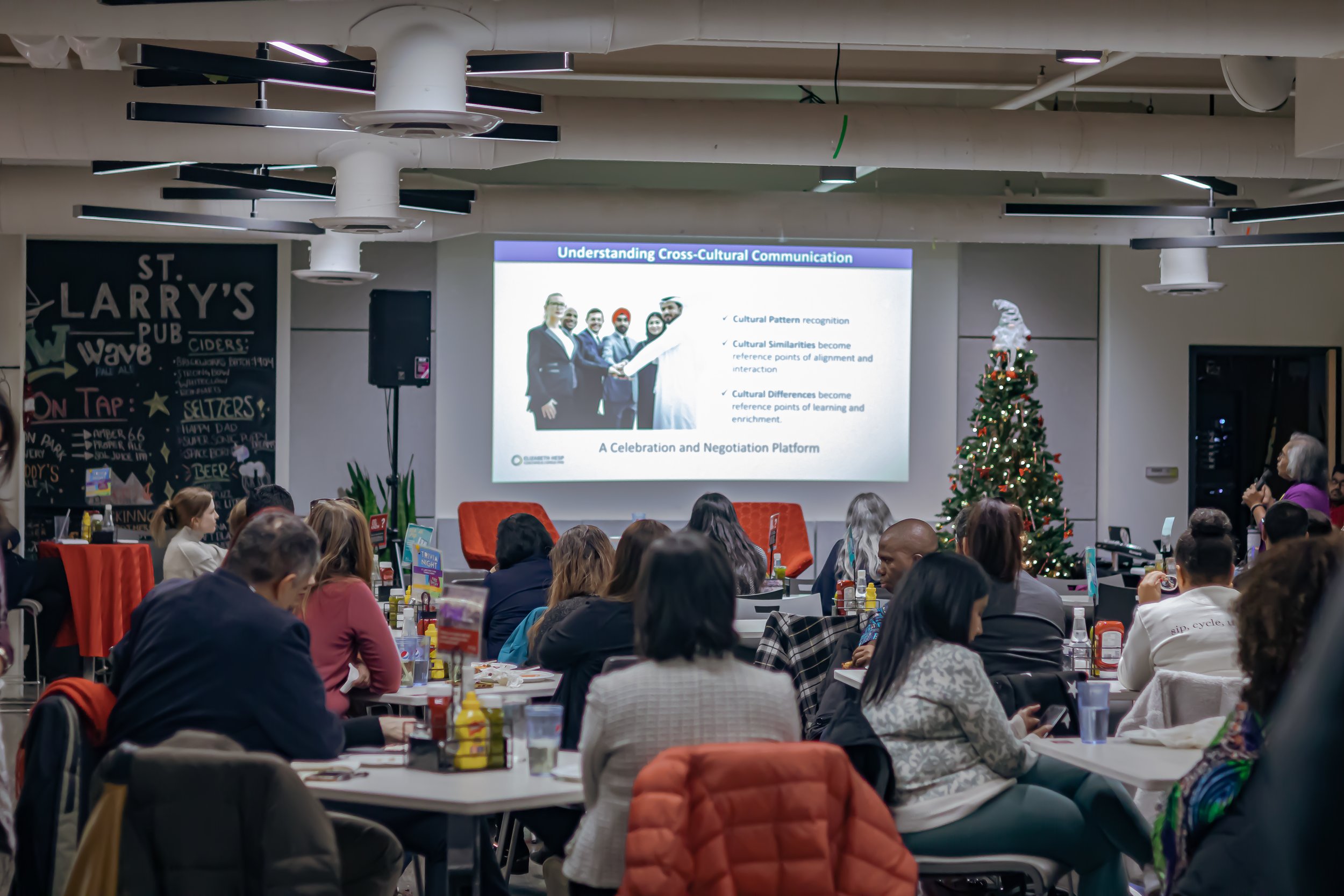 A conference or workshop in a decorated room with Christmas trees, Christmas lights, and Christmas ornaments. Attendees are seated at tables facing a large projection screen displaying a presentation about cross-cultural communication. The room has modern decor with a blackboard on the wall and some tables with snacks and drinks.