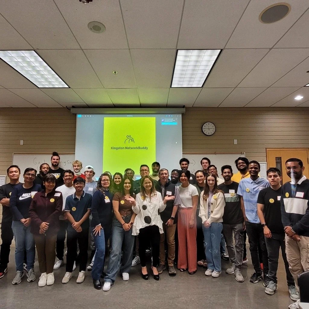 A group of diverse people posing for a photo in a classroom with a whiteboard and a projection screen displaying 'Kingston NetworkBuddy'.