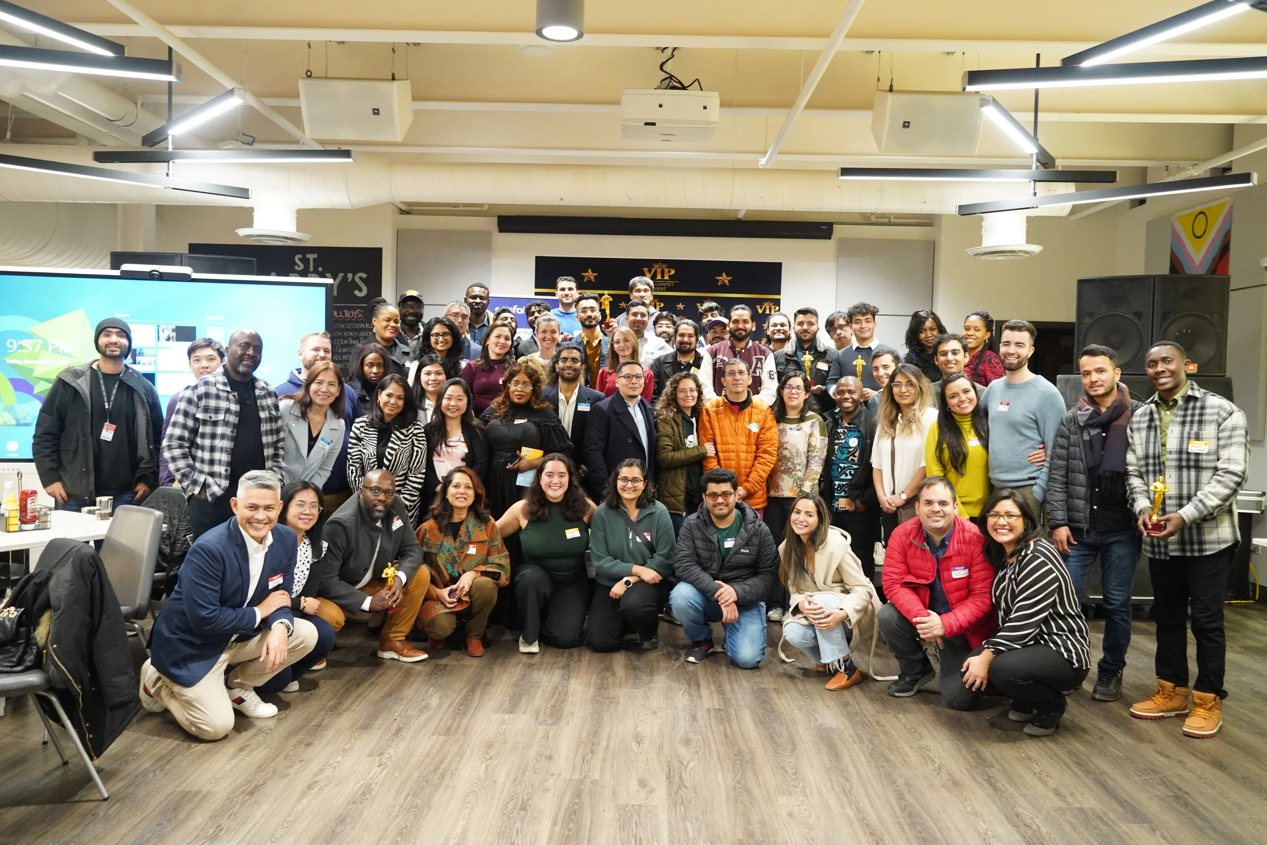 A diverse group of people gathered together for a photo at an indoor event or conference, with a presentation screen and banners in the background.