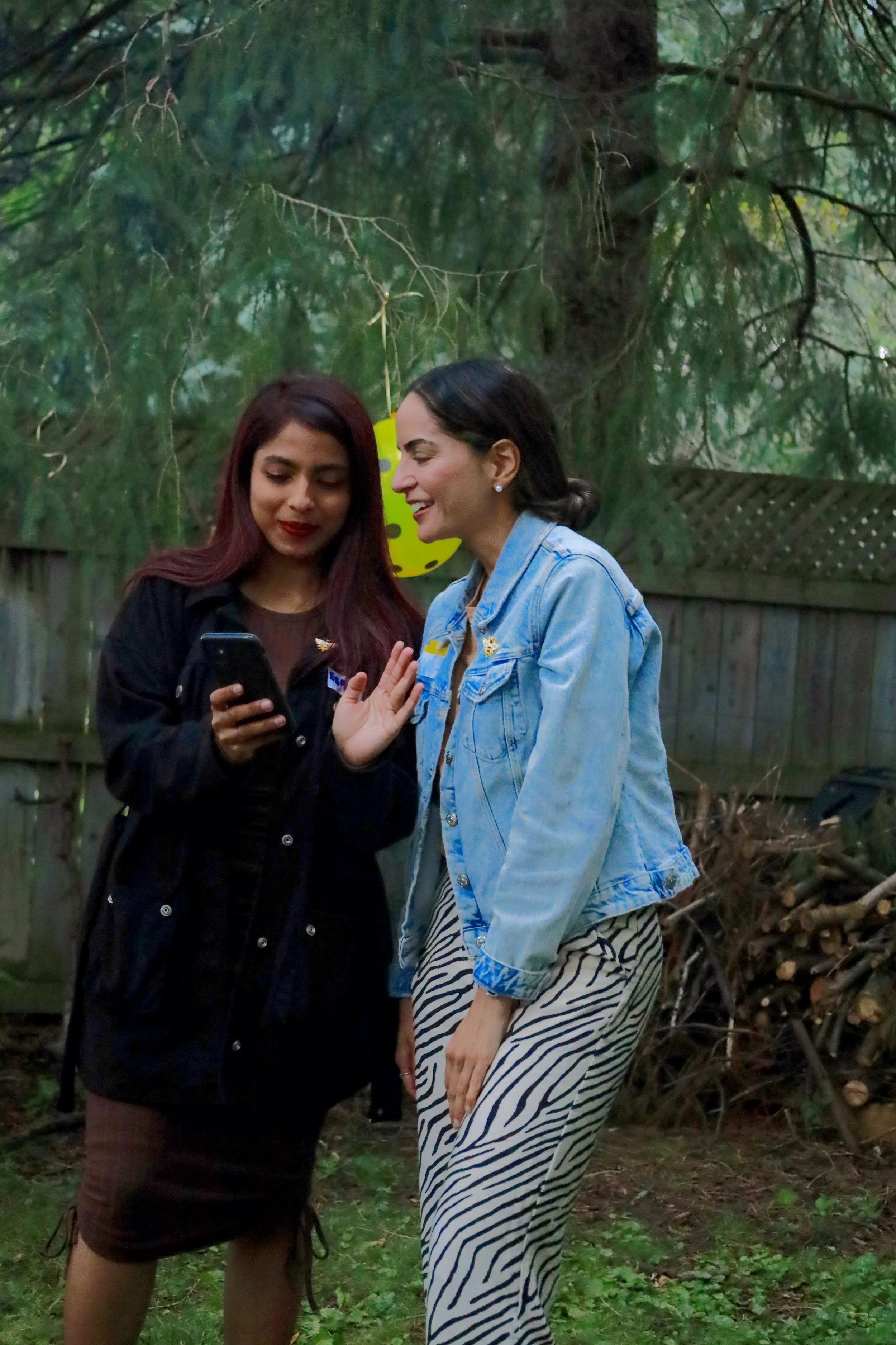 Two women are standing in a backyard, looking at a phone and sharing a laugh. One woman is wearing a black coat and a brown skirt, and the other is in a denim jacket and zebra striped pants. There is a yellow polka dot lantern hanging from a tree in the background.