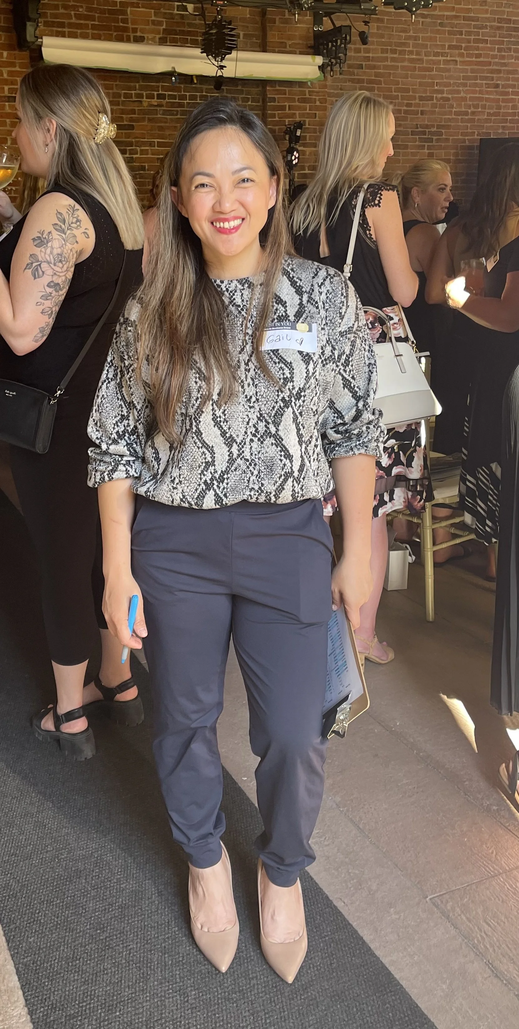 A woman with long brown hair, wearing a snakeskin patterned blouse, navy pants, and nude heels, smiling at a social gathering with other women in the background.