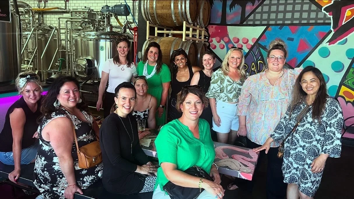 Group of women gathered in a brewery, some seated at a table and others standing, with brewing tanks and a colorful mural in the background.