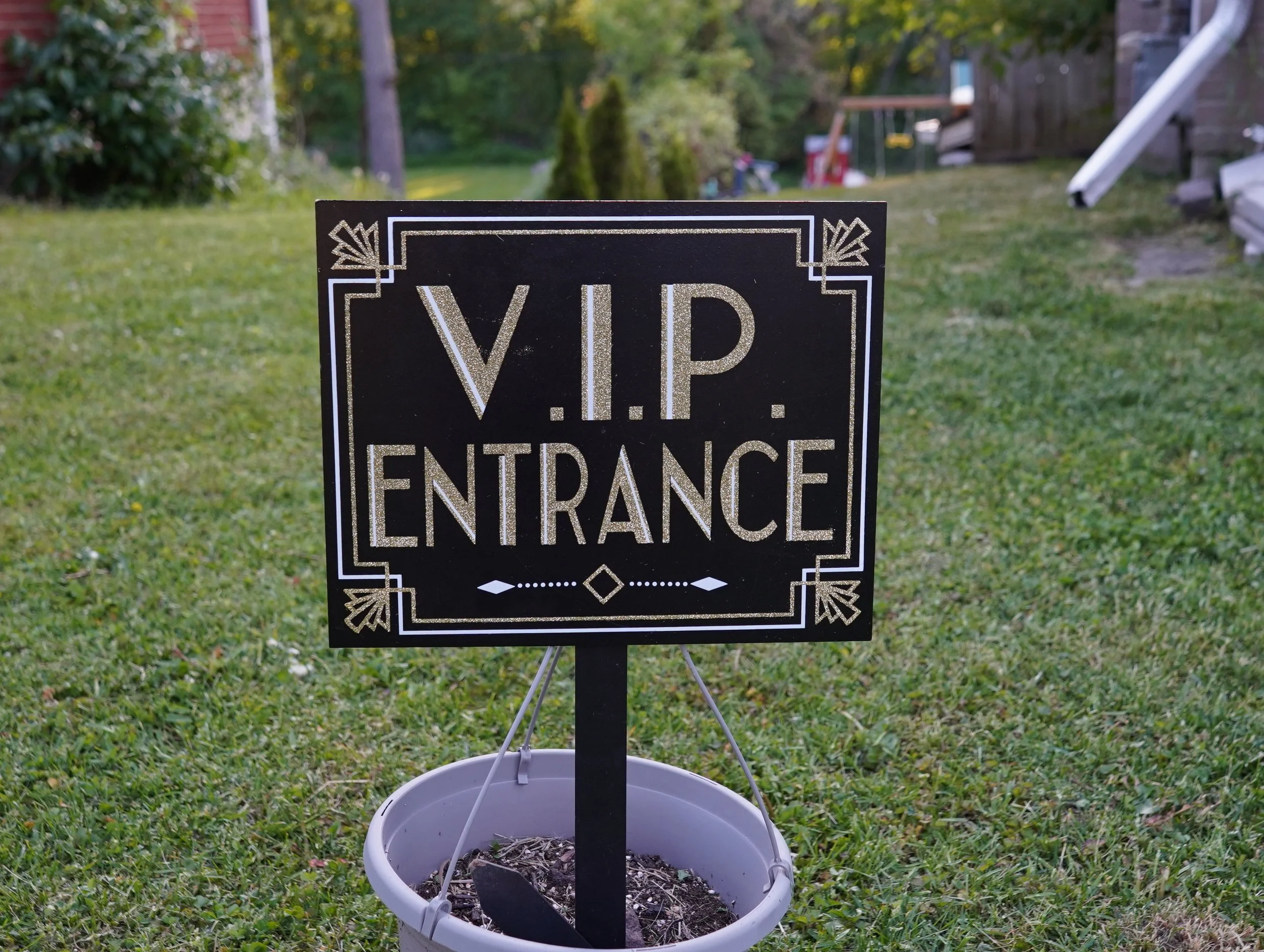 A black and gold sign with the words "V.I.P. ENTRANCE" in a decorative font, placed in a white bucket filled with soil in a backyard.