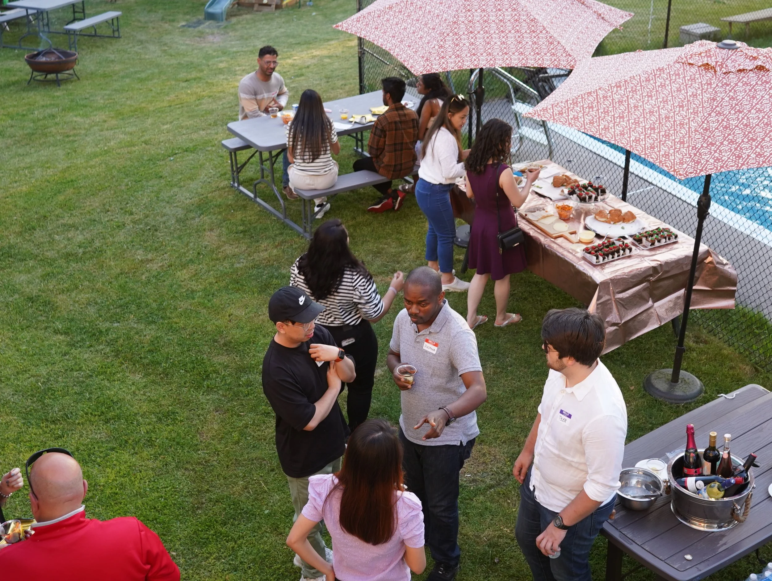 People having a backyard party with tables of food and drinks, a swimming pool nearby, and some umbrellas providing shade.