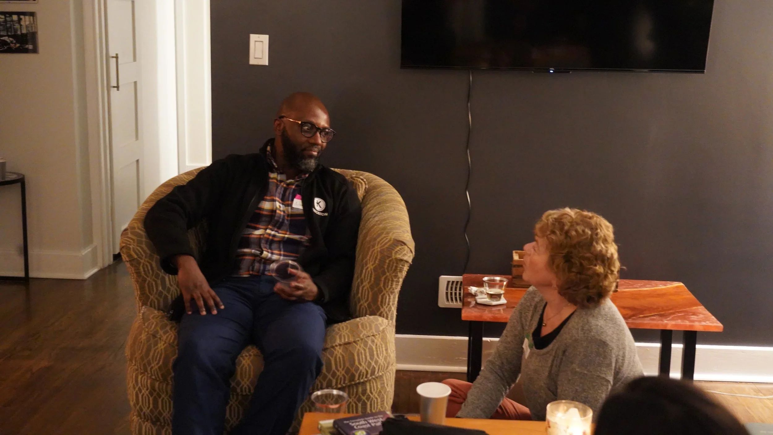 A man and a woman engaging in conversation in a cozy, dimly lit room. The man is sitting on a yellow patterned armchair, wearing glasses, a plaid shirt, and a dark jacket, holding a glass of wine. The woman is sitting on the floor, facing him, with curly blonde hair, wearing a gray sweater, and appears to be listening attentively. There are small tables with drinks and a candle in the foreground, behind the woman is a small table, and a large flat-screen TV is mounted on a dark wall.