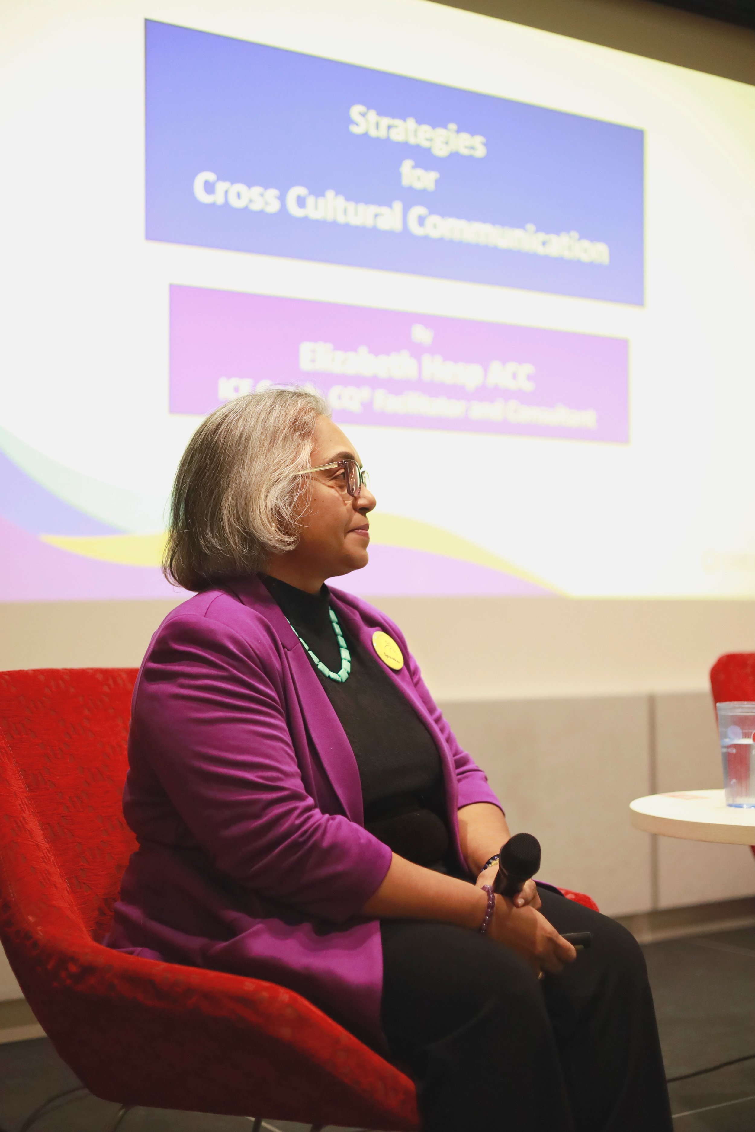 A woman with gray hair, glasses, wearing a purple blazer and a turquoise necklace, sitting in a red chair, holding a microphone during a presentation on cross-cultural communication.