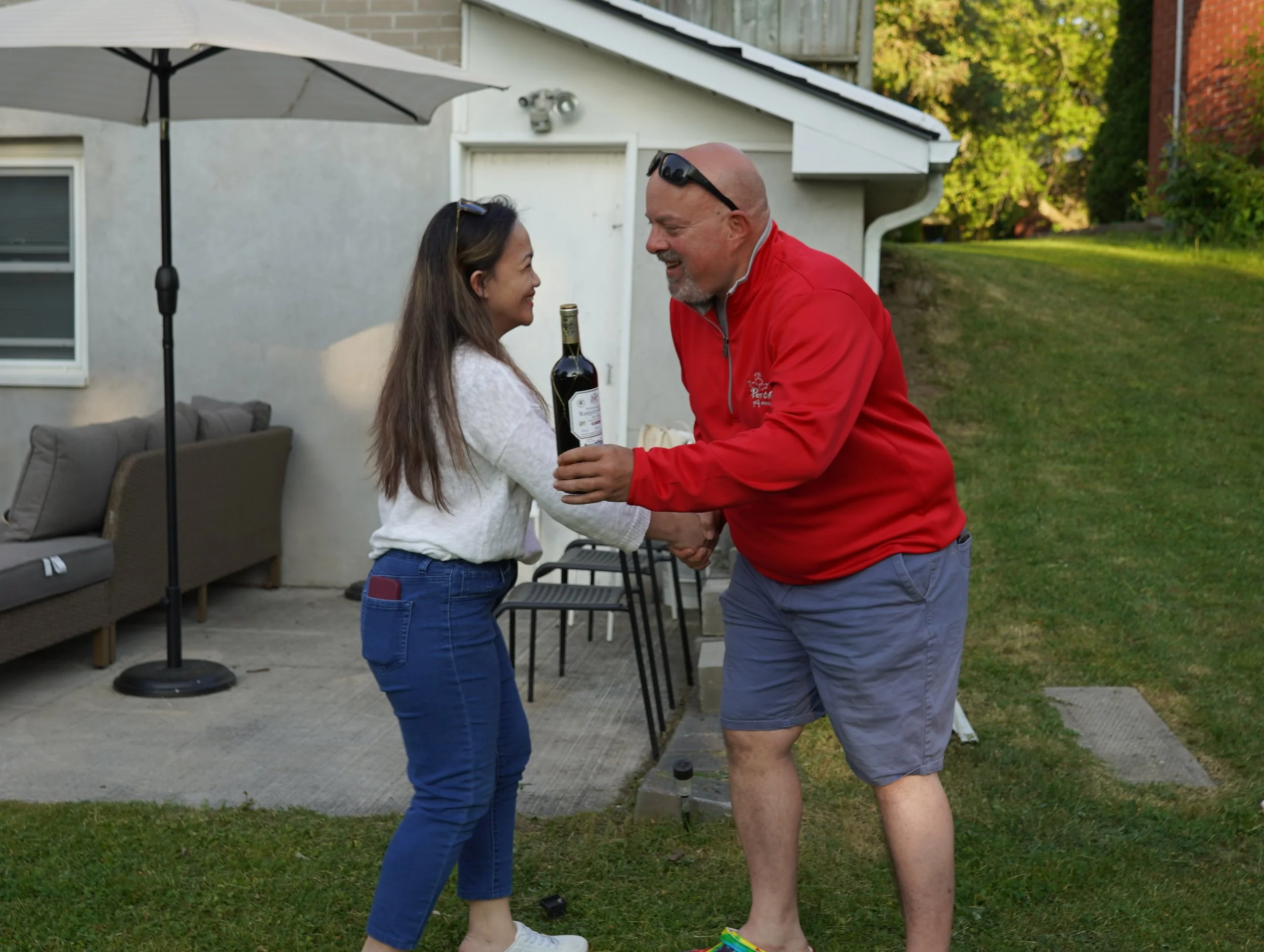 A man in a red jacket and gray shorts is giving a woman in a white sweater and blue jeans a handshake while holding a bottle of wine outdoors in a backyard.