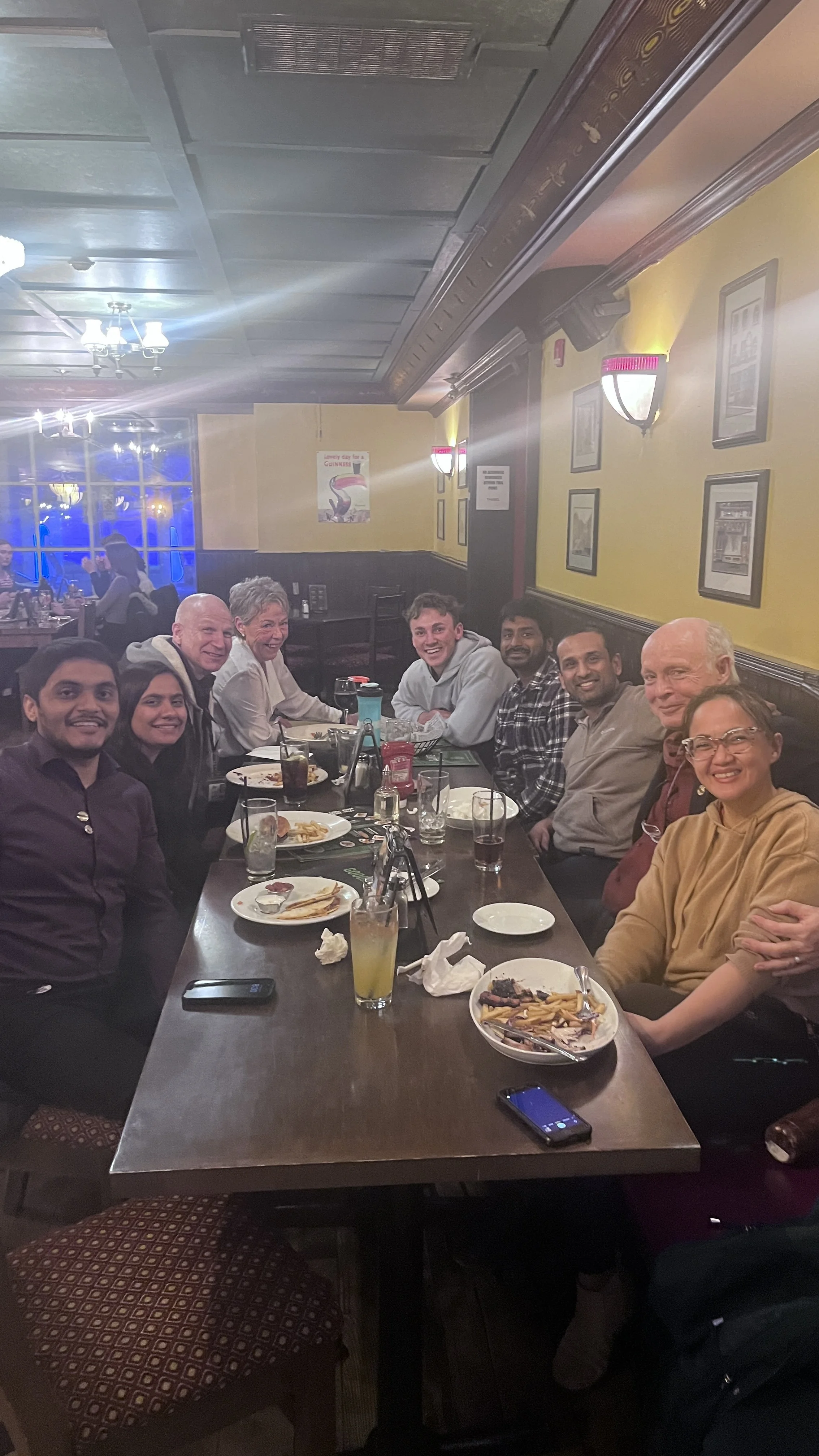 A group of nine people sitting around a long restaurant table with plates of food and drinks, smiling and posing for the photo