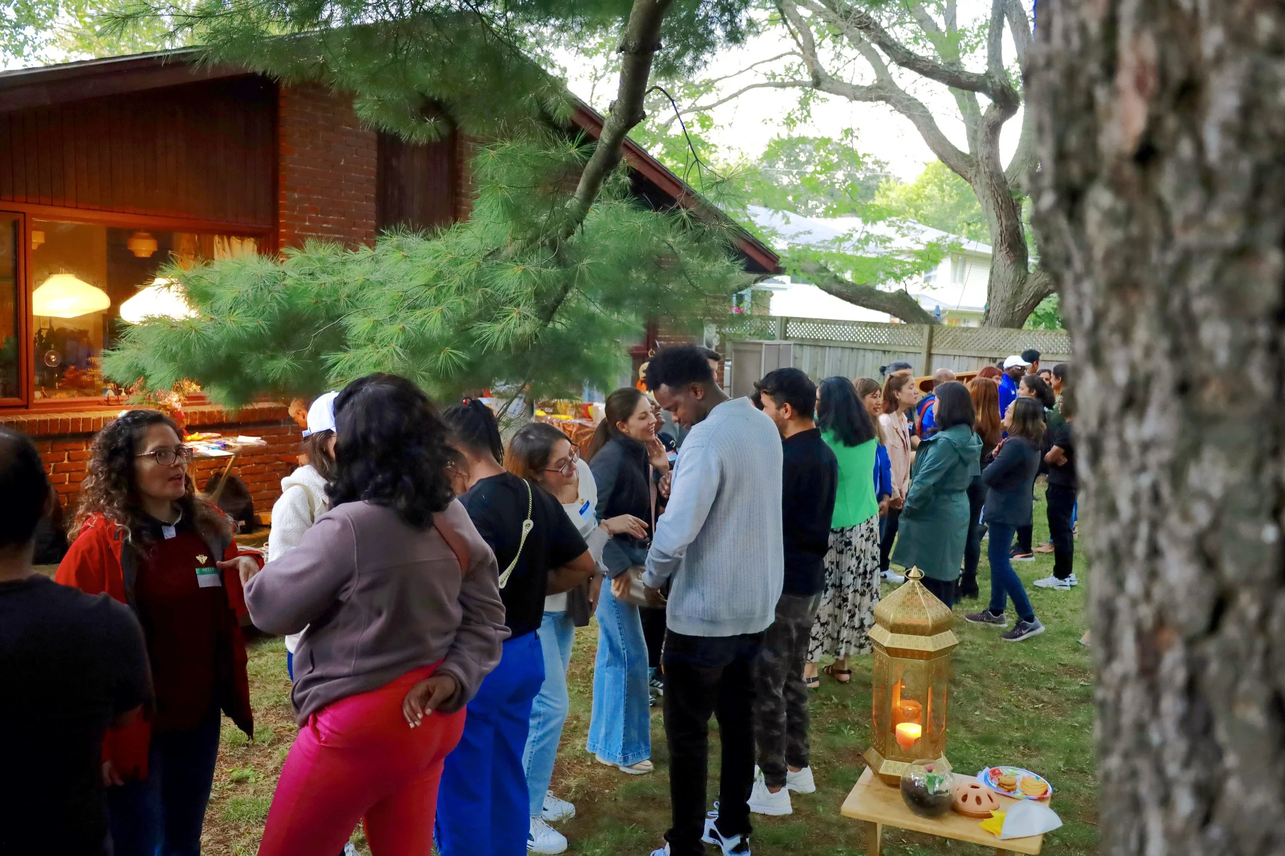 A group of people standing in a line outdoors during the evening, with some engaging in conversation. There are trees, a house with lit windows, and a table with a lantern, candles, and snacks nearby.