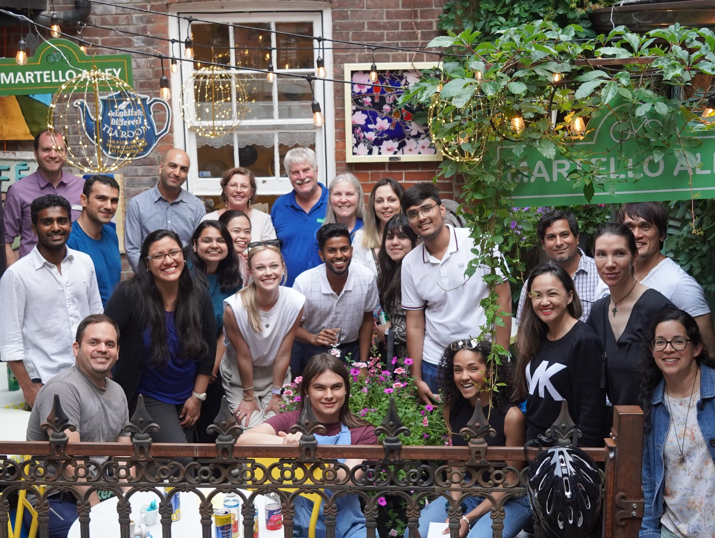 A large group of people gathered outdoors for a photo at a social event, with a brick building, decorations, and greenery in the background.