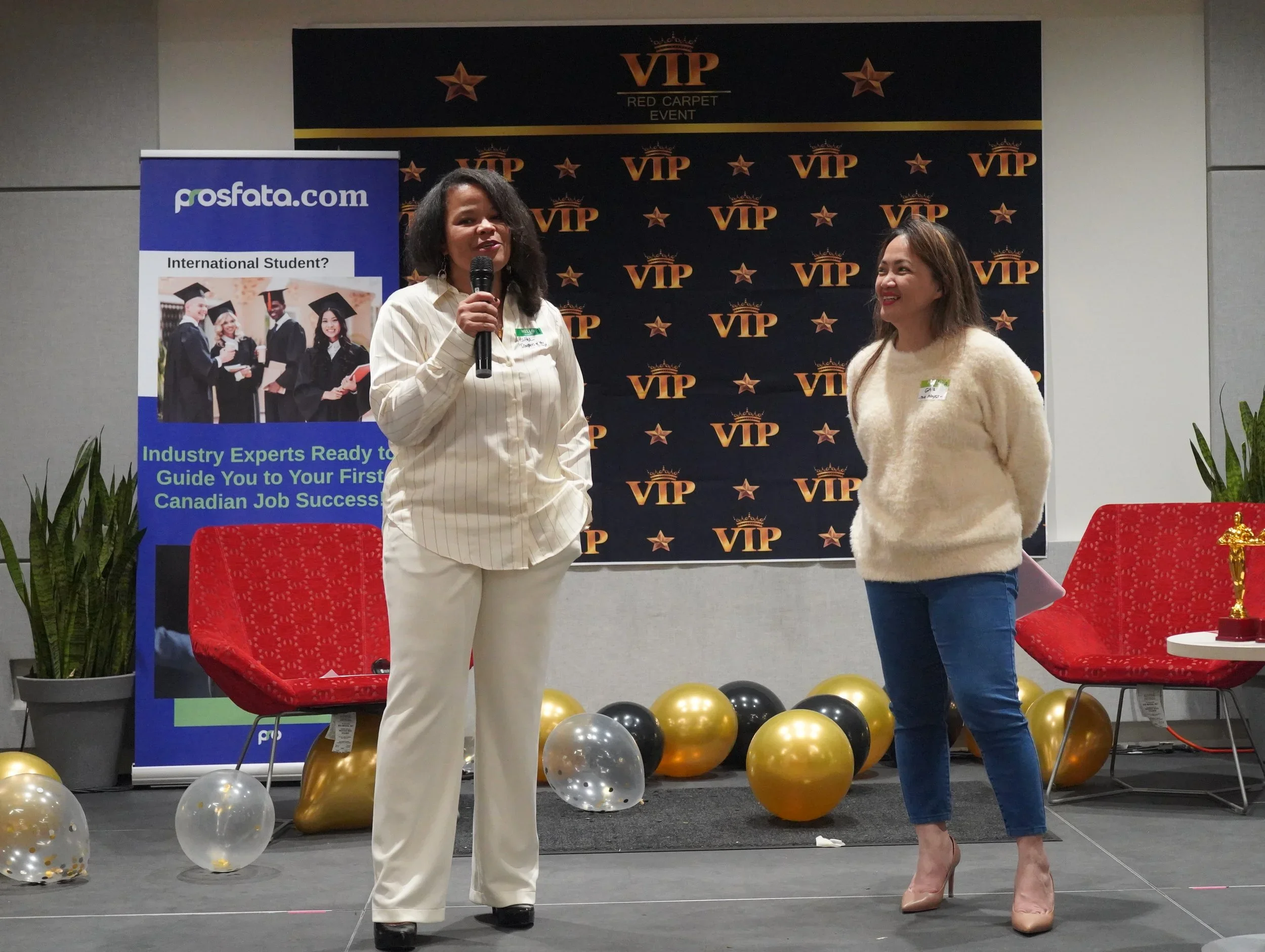 Two women standing on stage at an event. One woman is holding a microphone and speaking, the other is smiling and listening. Behind them are a VIP banner, balloons, a red chair, and a backdrop with VIP logos. To the left, a poster with people in graduation caps and gowns and a URL for prosfata.com.