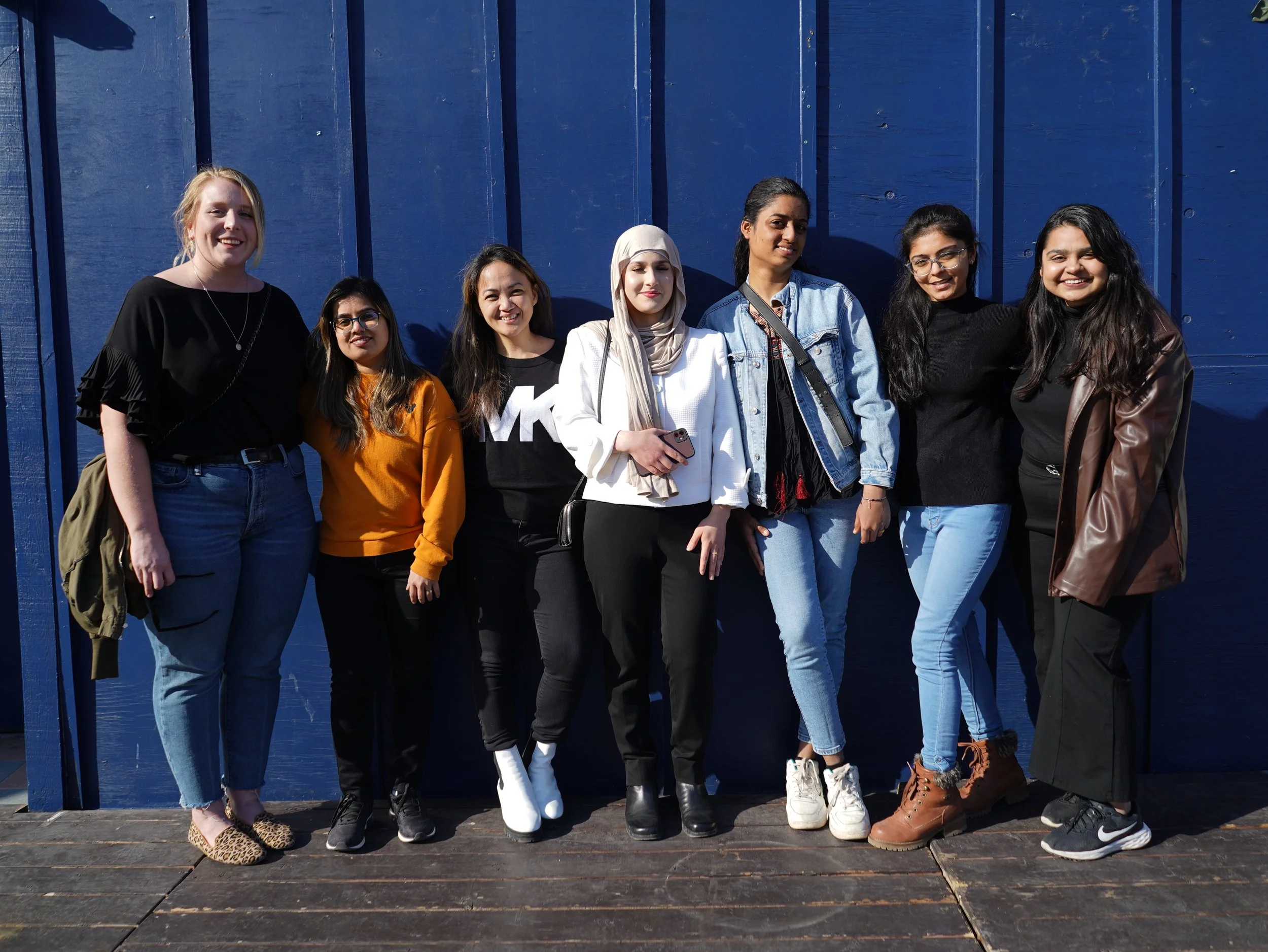 Group of eight diverse women standing together outdoors in front of a blue wooden wall, smiling at the camera.