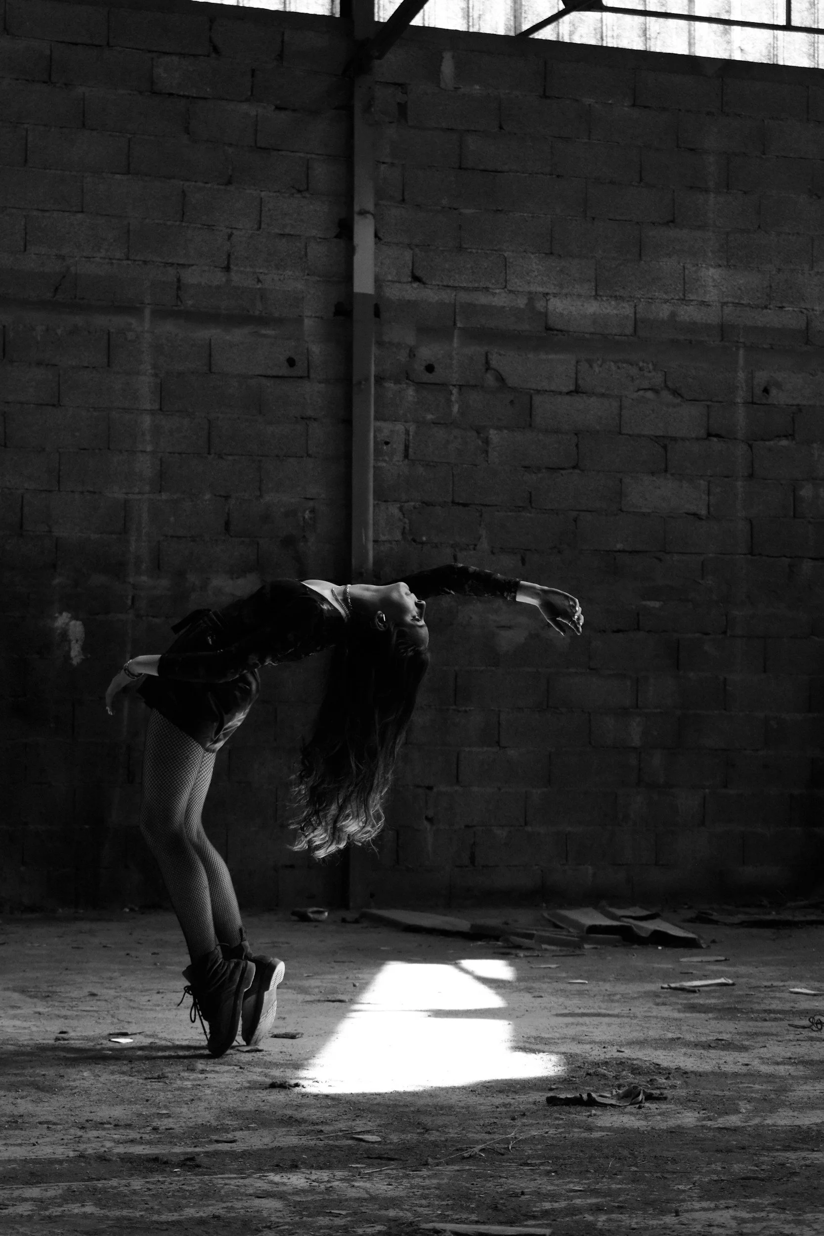 A woman with long hair performs a dance move in an industrial space with brick walls.