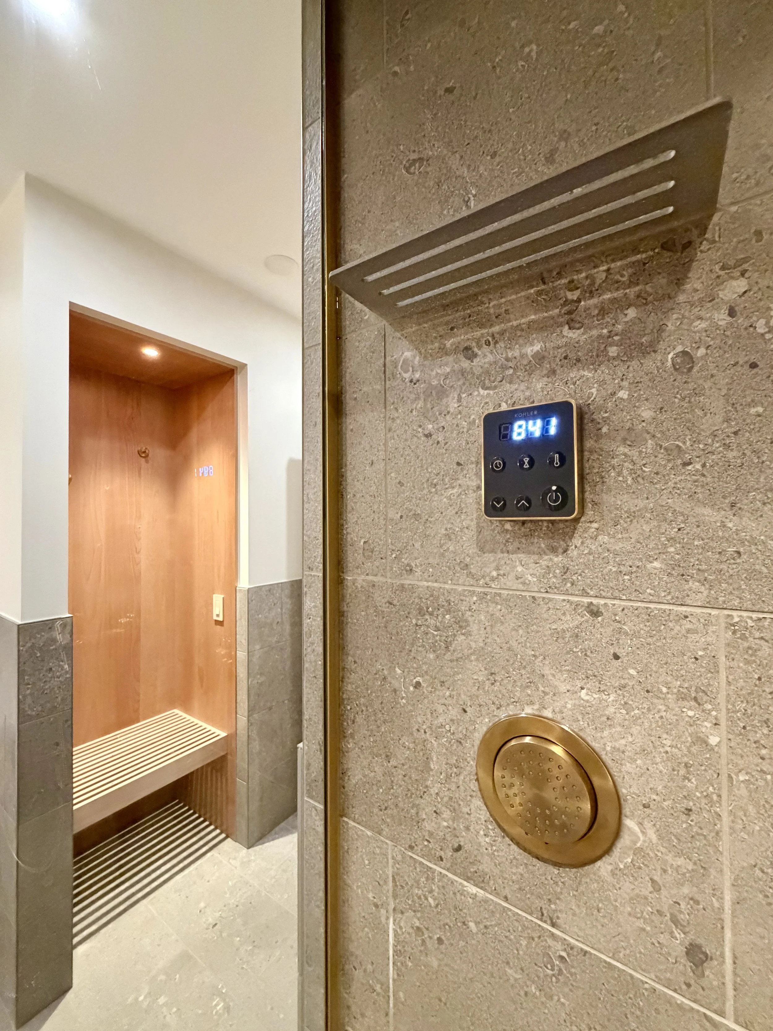 Foreground:

Architectural Details of Custom Built Steam Shower Including Schluter SHELF-W in Wave Greige, 1/2" Clear Glass & 1/2" Brushed Gold Structural Channel.

Steam Generator Control and Purist Body Spray in Vibrant Brushed Moderne Brass by Koh