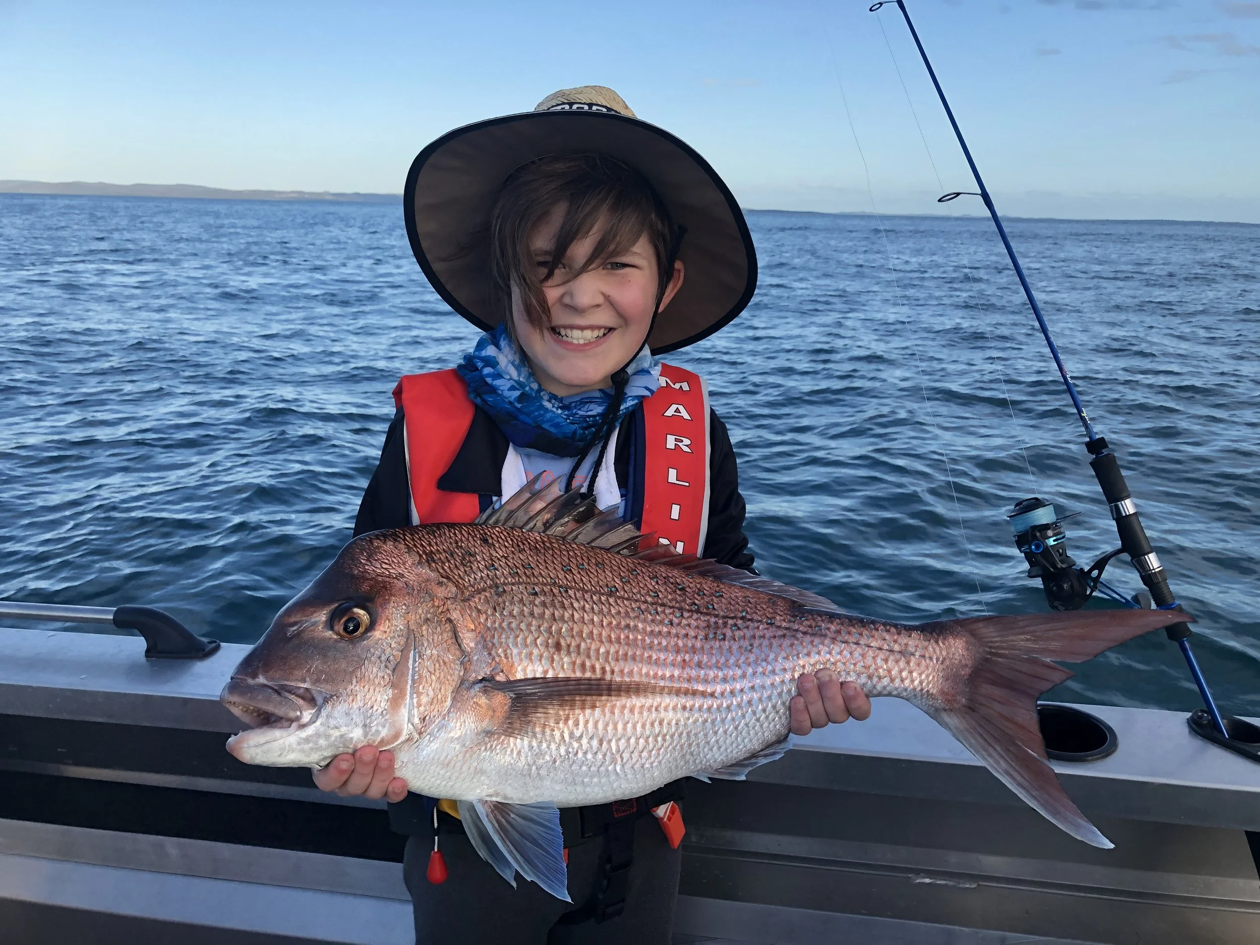 A young boy smiling and holding a large red snapper fish on a boat in the water. He is wearing a wide-brimmed hat, a red life jacket, and a blue neck gaiter, with a fishing rod on the boat.