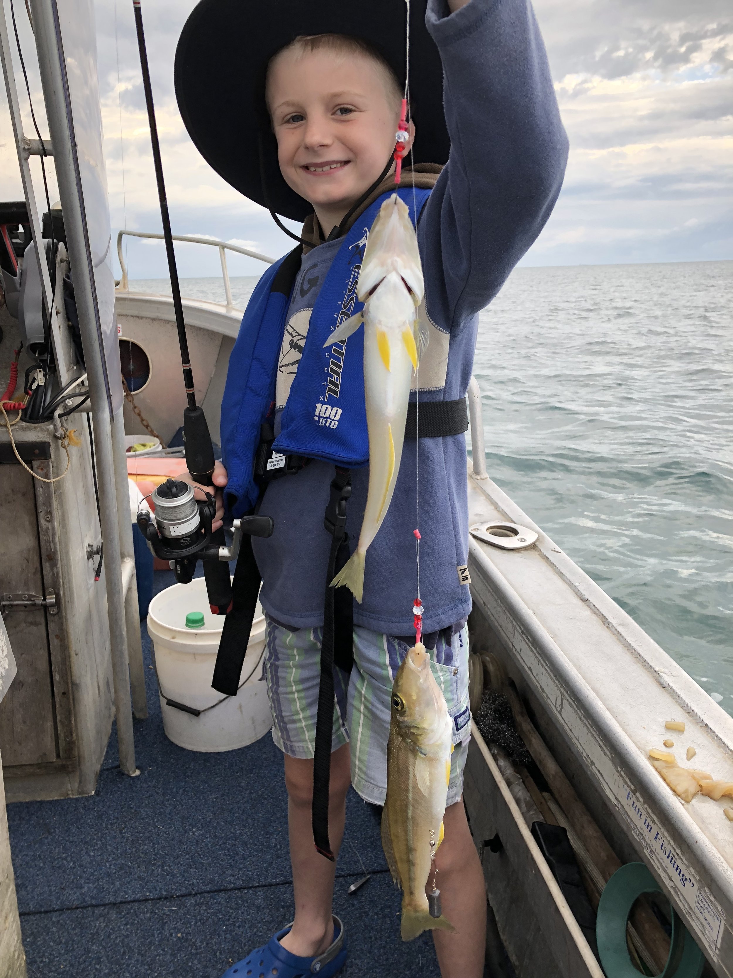 A young boy on a boat holding up a fishing line with two fish caught, smiling at the camera. The sea and sky are visible in the background.