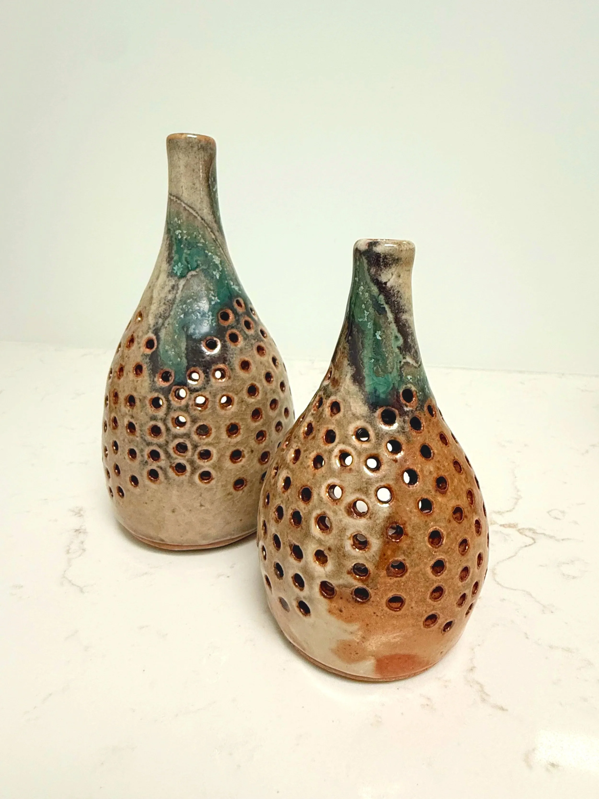 Two ceramic vases with holes and glazed in earthy tones, featuring bluish-green accents, placed on a white marble surface.