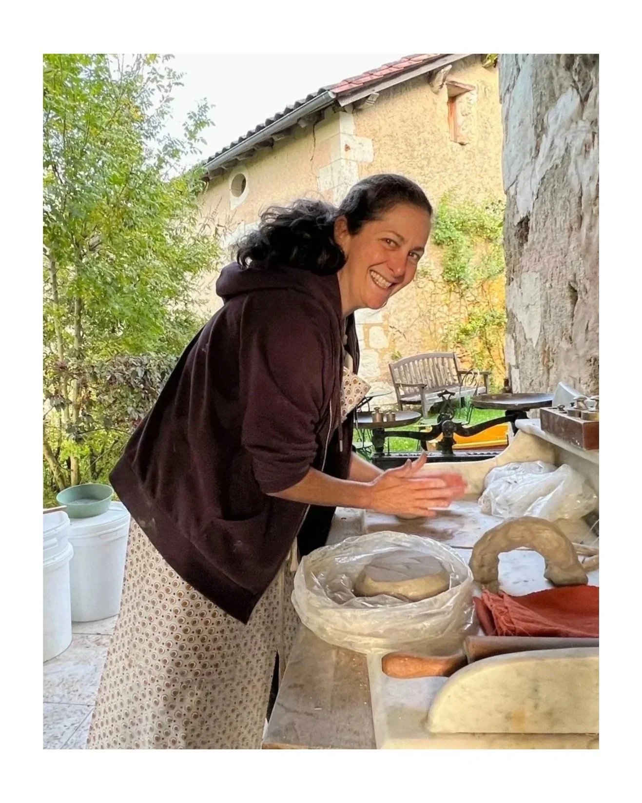 A woman baking bread outdoors, smiling, with her hands covered in dough, surrounded by baking tools and ingredients on a counter.